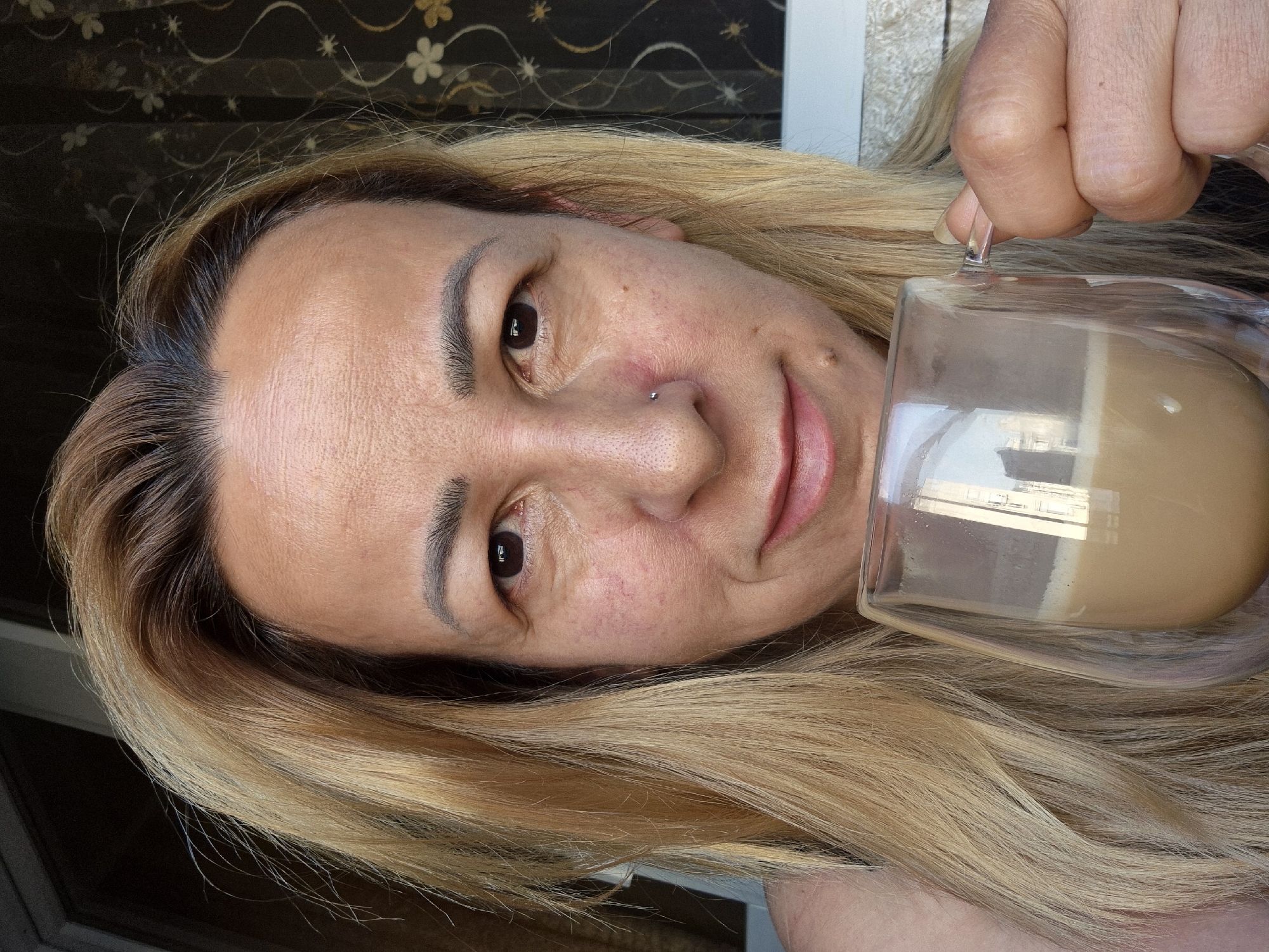 A medium close-up selfie shows a woman with blonde hair holding a transparent double-walled glass mug filled with a light brown beverage. The woman is looking directly at the camera with a slight smile. She has a small nose piercing. Her skin shows some redness and texture. Behind her, a dark curtain with a floral pattern is visible. The lighting suggests an outdoor setting, possibly a balcony or patio. The background is slightly out of focus, but a railing and part of a building with windows can be seen reflected in the mug. The image was taken in the city of Unknown, Unknown.