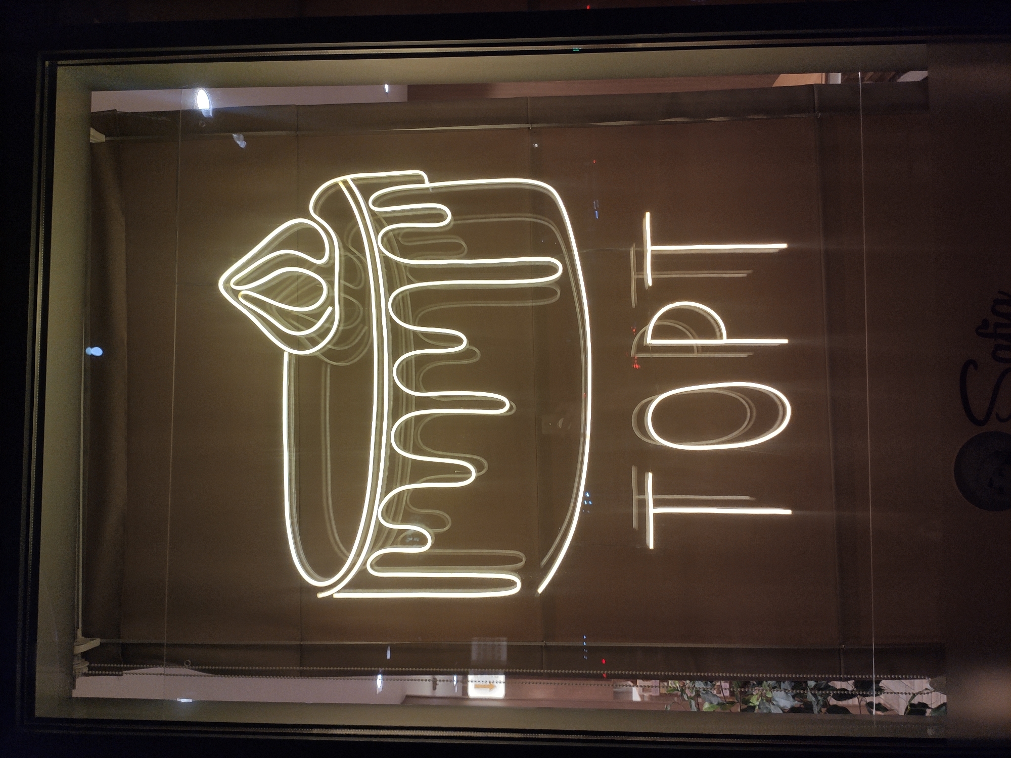 The image shows a neon sign in a shop window. The sign depicts a stylized cake with frosting dripping down the sides, and below it, the word "TOPT" is written in neon letters. The background is a brown wall, and a roller blind is visible behind the sign. To the left of the sign, a window reveals a dimly lit interior with plants and what appears to be a store or cafe. The image is taken at night, as indicated by the reflections in the glass and the darkness outside. The overall impression is of a bakery or dessert shop advertising its services. It is located in Tashkent, Uzbekistan.