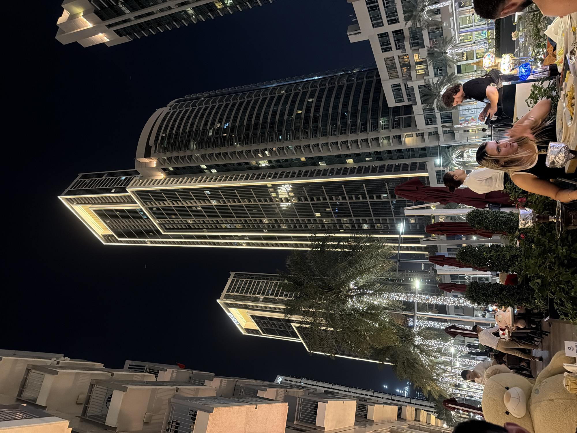 A view of downtown Dubai at night shows a high-rise building and a lit tree. Buildings are illuminated with white lights, and the night sky is black. People are present in the scene.