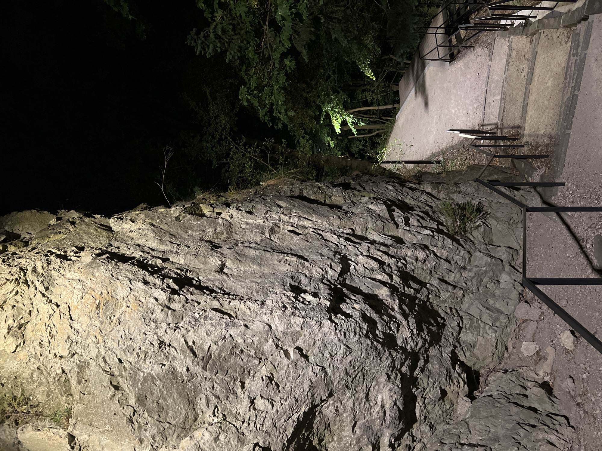 The image depicts a stone staircase descending beside a large rock face at night. The rock face, illuminated by artificial light, occupies the left side of the frame and shows rough, textured surfaces. The staircase, made of concrete steps with stone edges, is lined with black metal railings. To the right of the stairs, a pathway curves away into a dark wooded area, also lit by artificial lights that cast a green glow on the foliage. The overall scene is one of an outdoor walkway or trail, likely in a park or natural setting, that is open for nighttime use. The specific location in Nitra, Slovakia is the Zobor hill, specifically the trail leading to the Zobor monastery ruins.