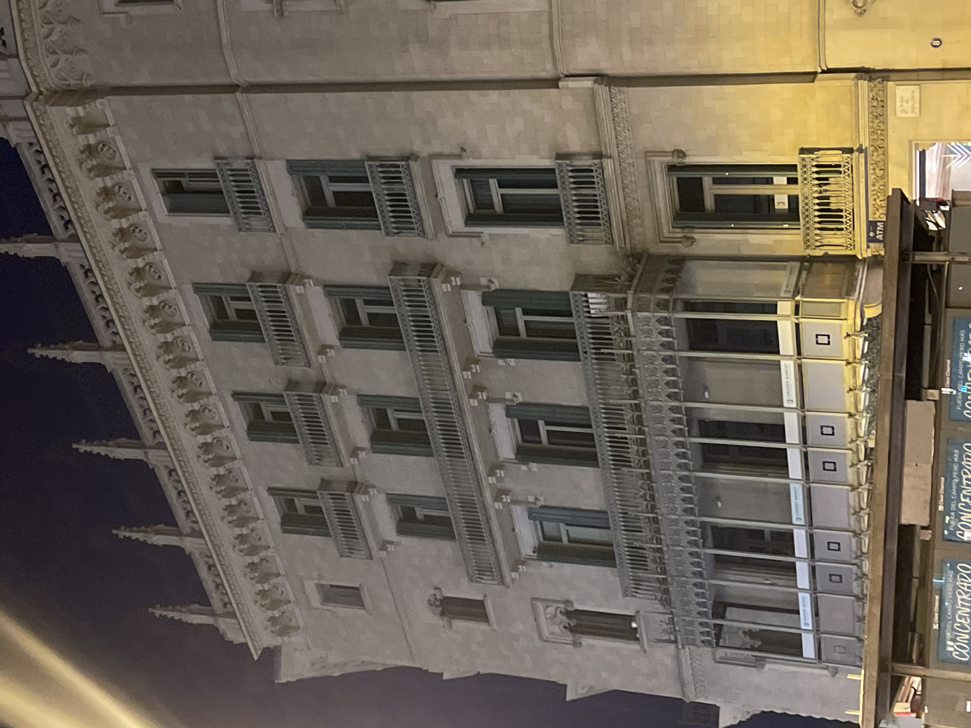 The image shows the facade of a grand building at night in Barcelona, Spain. The building is made of stone and features multiple stories with regularly spaced windows and balconies. The architectural style appears to be neo-Gothic or neo-classical, characterized by ornate detailing, decorative carvings along the cornices, and pointed finials at the top.
Each floor has a series of windows, many of which have green shutters that are partially open. Several balconies with decorative railings extend from the upper floors, adding to the building's elaborate appearance. The lower section of the building has a more modern section with large glass panels and signage. Below this, a street-level structure with billboards is visible. One billboard prominently displays the word "CONCENTRADo" in large letters, suggesting an advertisement.
The lighting indicates it is nighttime, with artificial light illuminating the facade and the street level. A bright, blurred light streak is visible in the upper left corner, possibly from a streetlamp or vehicle headlights. The sky above is dark. An ATM machine is visible on the right side of the building, near street level. The overall impression is one of a well-maintained, historic urban building.