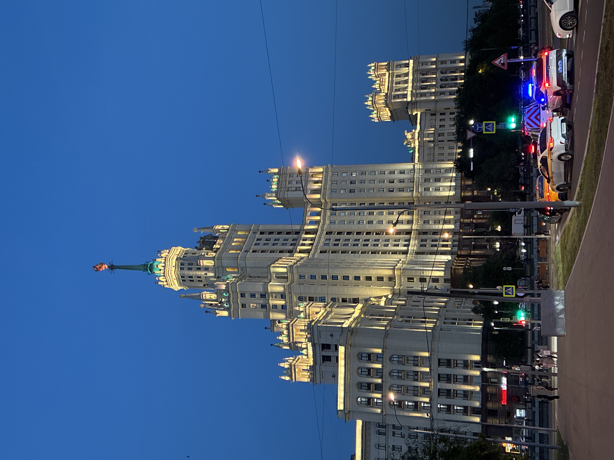 The image displays an illuminated Stalinist skyscraper in Moscow, Russia, under a dark blue evening sky. The grand, multi-tiered building, likely one of the city's "Seven Sisters" (specifically the Kotelnicheskaya Embankment Building), is brightly lit with warm yellow lights that accentuate its intricate architectural details and tall spire, which is topped with a star-like ornament.
In the foreground, a busy street scene unfolds. Several vehicles are present, including an emergency vehicle with flashing blue lights and white and yellow cars, some possibly taxis. Traffic lights display both green and red signals. Various road signs are visible, including a yellow diamond-shaped sign with a black border and a pedestrian crossing sign. A few pedestrians can be seen on the sidewalk. Overhead wires stretch across the sky in places.
The scene depicts urban activity during dusk or night, characterized by the dark sky and extensive artificial lighting from the building, streetlights, and vehicle lights. The weather appears clear. Faint Cyrillic text is visible on a red sign at street level, possibly "Московия". The overall atmosphere is one of a vibrant and well-lit city night, highlighting an iconic Moscow landmark.