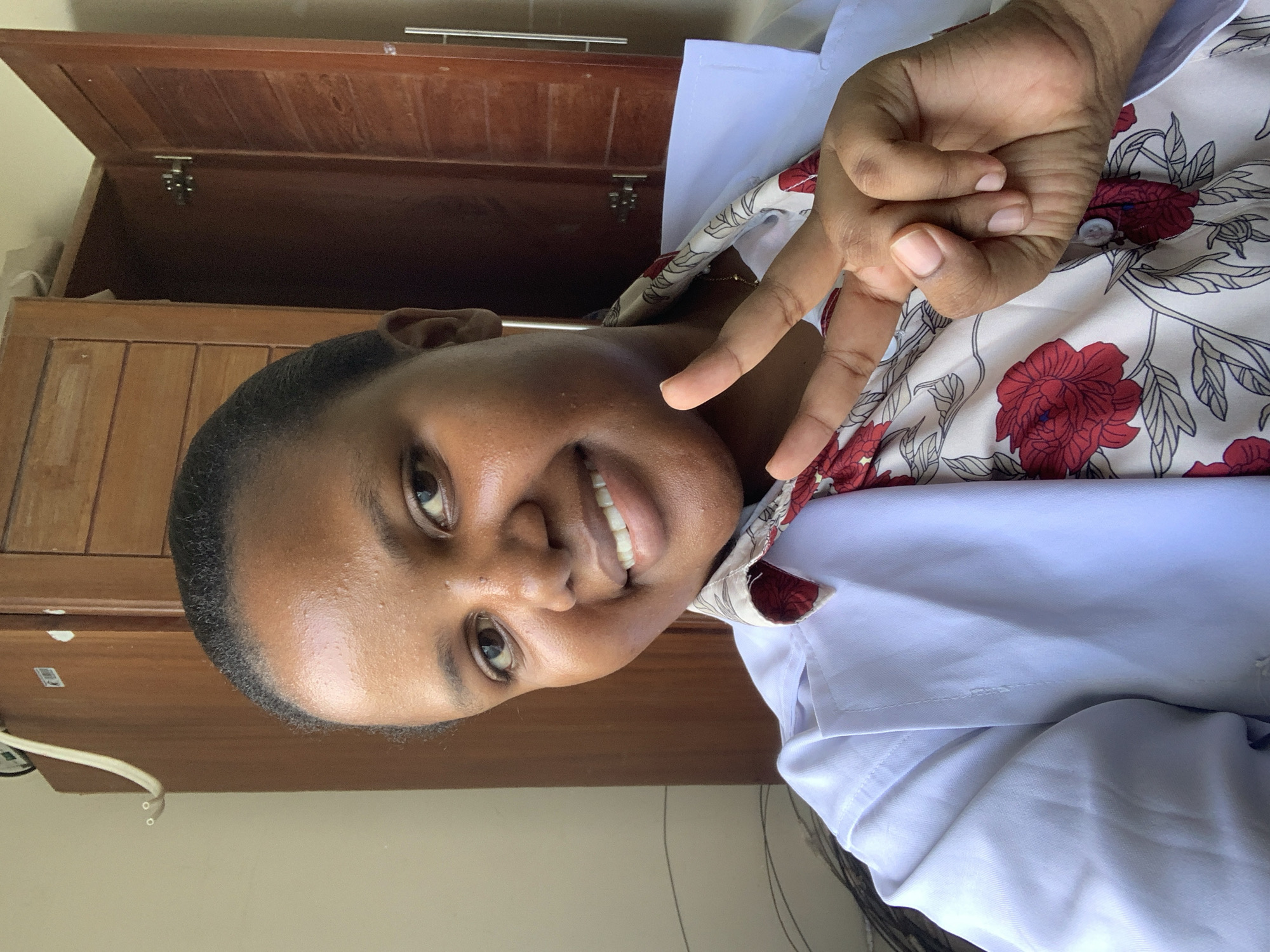 A smiling woman is making a peace sign with her right hand in front of a wooden cabinet. She is wearing a white top with a floral pattern on the collar. The cabinet is made of dark brown wood. The wall is light beige. Location information is unavailable.