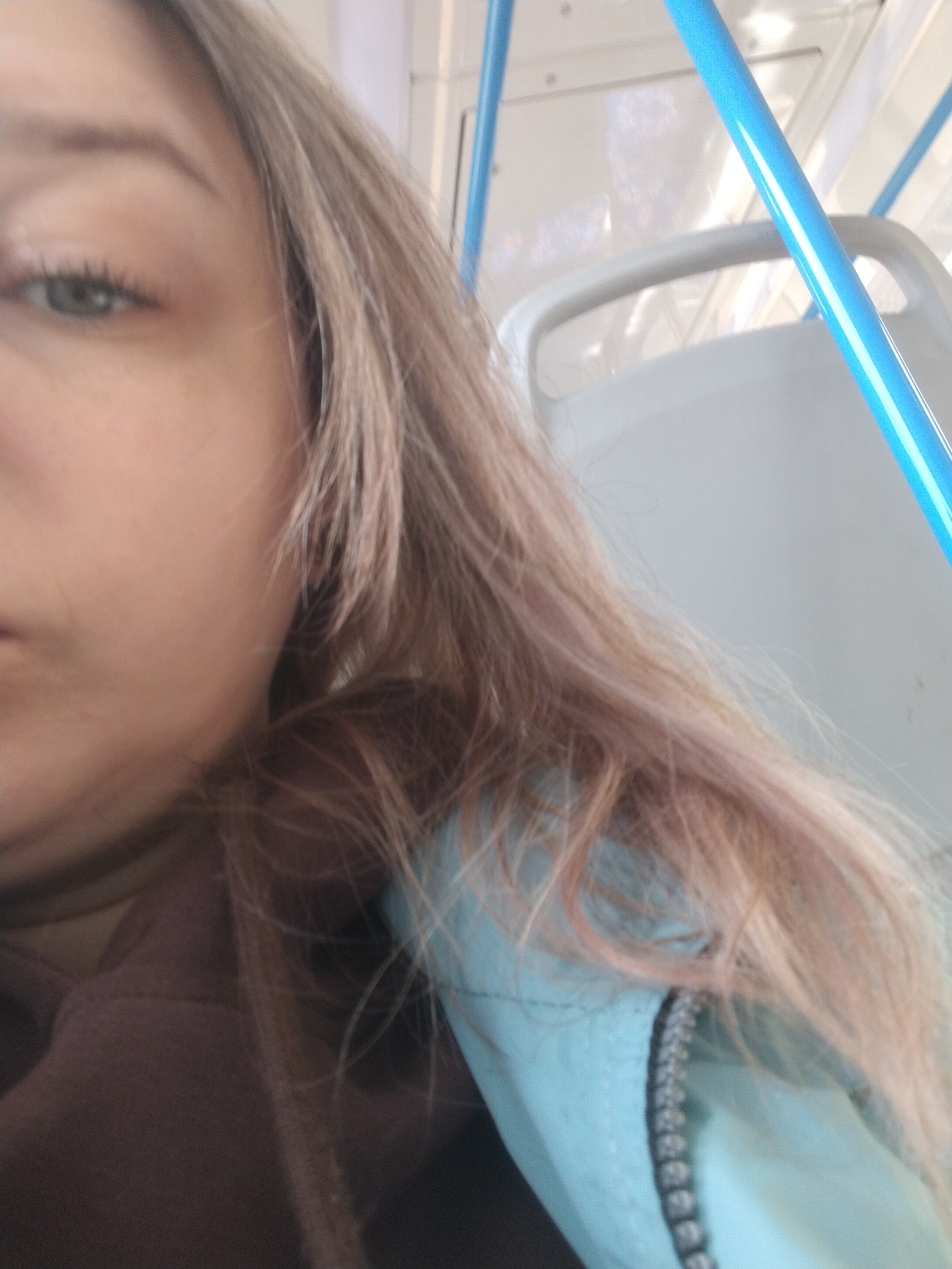 A close-up indoor shot features a person with fair skin and light brown hair with hints of pink. Only a portion of their face is visible, including one eye with green iris, part of their cheek, and a hint of their mouth. They are wearing a dark brown top. Adjacent to their hair, a light blue jacket with a silver zipper is seen. In the background, the interior of a public transportation vehicle is visible. There is a gray seat with a handle, and a blue pole, and the overhead area is white with what appears to be a window. The location is potentially in Rusanovo, Russia.