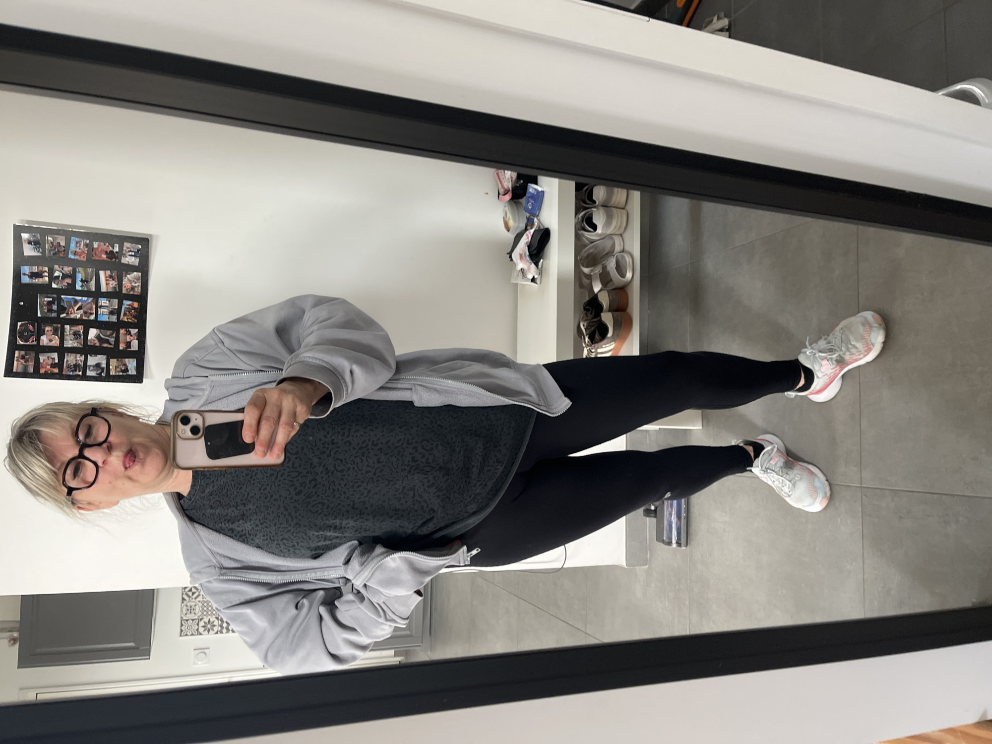 A middle-aged woman is taking a selfie in a mirror. She is wearing glasses, a gray t-shirt with a subtle pattern, black leggings, a gray zip-up jacket, and pink and gray running shoes. She is holding a smartphone in her right hand.
The reflection shows the interior of a room. Behind her, there is a white wall with a collage of photos on it. A shoe rack with various shoes is visible. The floor is gray tile.
The location is Chambœuf, France.