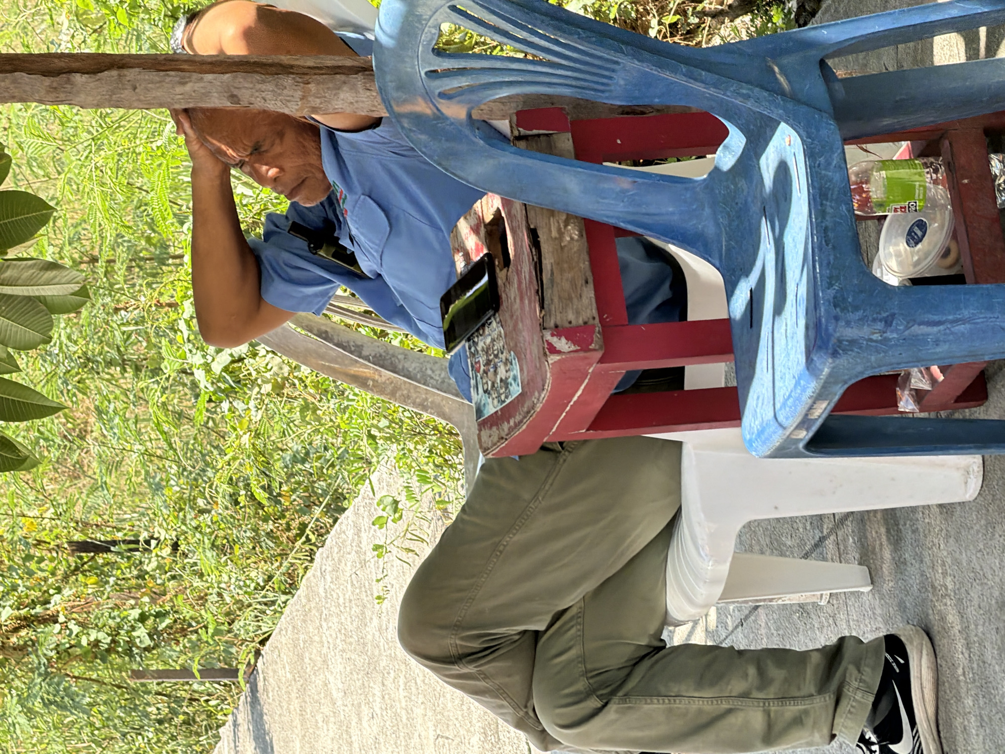 An elderly man in a blue collared shirt and green pants is seated on a white plastic chair. His legs are crossed, and his right hand is resting on his head. He appears to be dozing or resting. Next to him, on a small red and brown wooden table, is a black mobile phone. A blue plastic chair is positioned in front of the man, partially obscuring him. The scene appears to be outdoors, with lush green foliage and trees in the background. Sunlight filters through the leaves, casting shadows. The ground beneath the chairs is paved with stone. A metal post is visible to the left, and a wooden post is directly behind the man. The environment suggests a relaxed, possibly casual setting, perhaps in a garden or an outdoor seating area in Ko Samui District, Thailand, given the tropical vegetation.