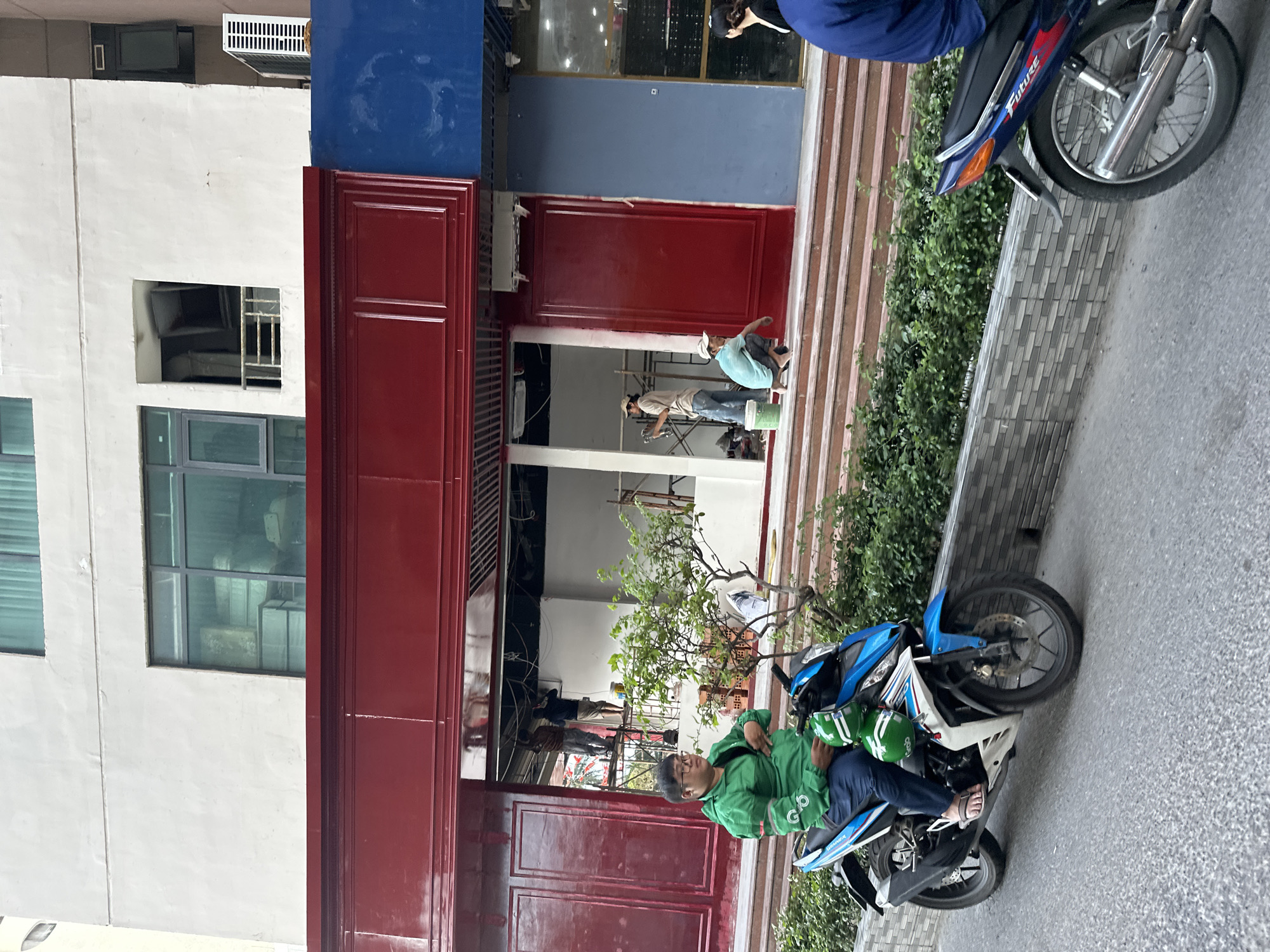 The scene shows a man wearing a green jacket and glasses sitting on a blue and white motorcycle parked on a street in Bac Nha Trang Ward, Vietnam. Two green helmets sit on the motorcycle's seat. The man is looking towards the building behind him. In the background, two individuals are working on the facade of a building. One person is seated and appears to be applying paint. Scaffolding is set up inside the building. The building's facade is being renovated, with red and blue paint visible. The street slopes downward towards the right. A bush is planted between the building and the sidewalk. Another motorcycle is visible on the upper right side of the image. The image is slightly rotated.