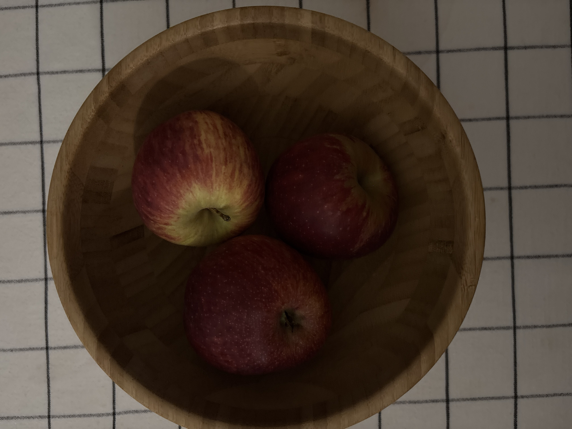 A high-angle close-up shot features three red apples in a wooden bowl placed on a checkered cloth. The apples are a mix of red and yellow hues. The bowl's wooden texture is visible. The checkered cloth underneath is white with dark gray lines. The overall lighting is somewhat dim, casting shadows within the bowl. The location and city, Saint Petersburg, Russia, are not identifiable within this visual information.