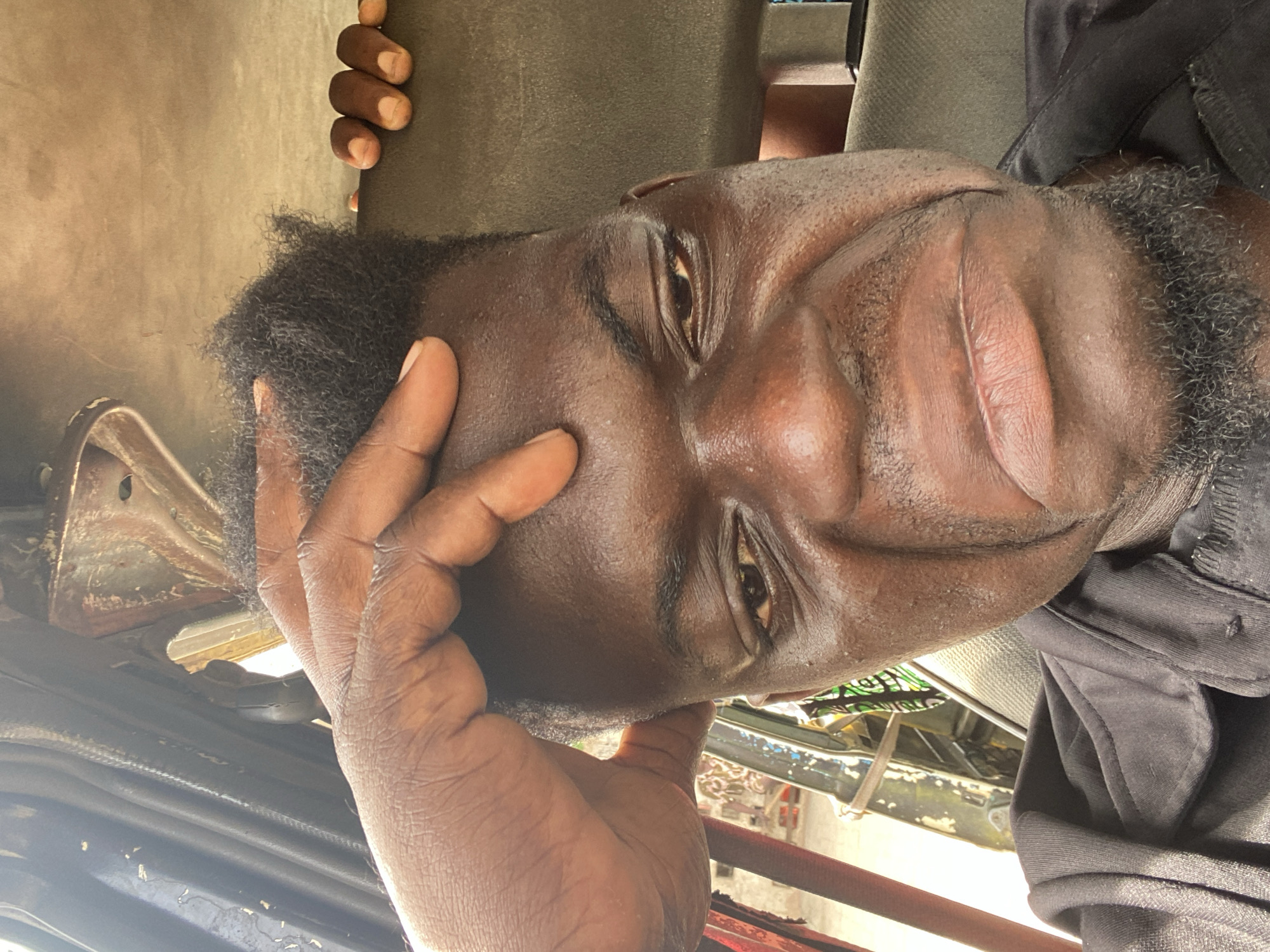The image is a close-up portrait of a Black man, presented with a 90-degree clockwise rotation. When viewed upright, the man is looking directly at the camera with a neutral to slightly pensive expression. He has a dark complexion, short curly black hair, and a short beard or stubble. His left hand is raised, with fingers resting against his temple and forehead. A portion of his right hand is visible in the upper right corner, seemingly resting on a surface.
He is wearing a dark, possibly black, long-sleeved garment, which appears to be a shirt or jacket. His skin shows a noticeable sheen, possibly from sweat or oil.
The background suggests an interior setting, likely within a vehicle such as a bus or car. Behind the man, there is a dark, possibly fabric-covered surface, consistent with a seat or seatback. To his left (when upright), bright natural light streams in from an opening or window, illuminating parts of his face. Through this opening, some indistinct metal structures and what appears to be a hint of a green patterned object or fabric are visible.
The lighting indicates it is daytime. There is no visible text in the image.