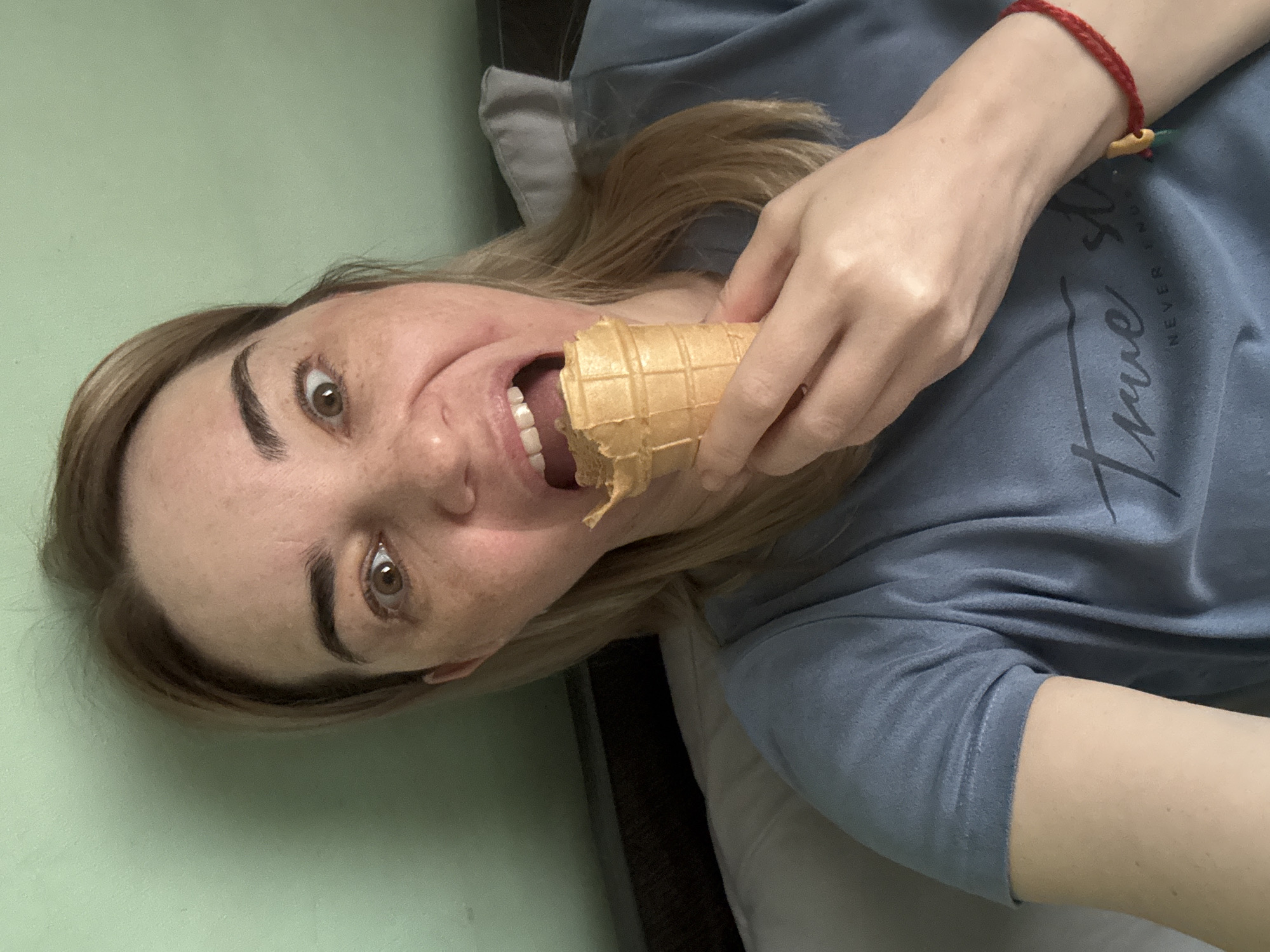 A woman is lying down eating an ice cream cone. She is wearing a blue t-shirt and a red bracelet. She is indoors and lying in what appears to be her bed. The location of the scene is Unknown, Unknown.