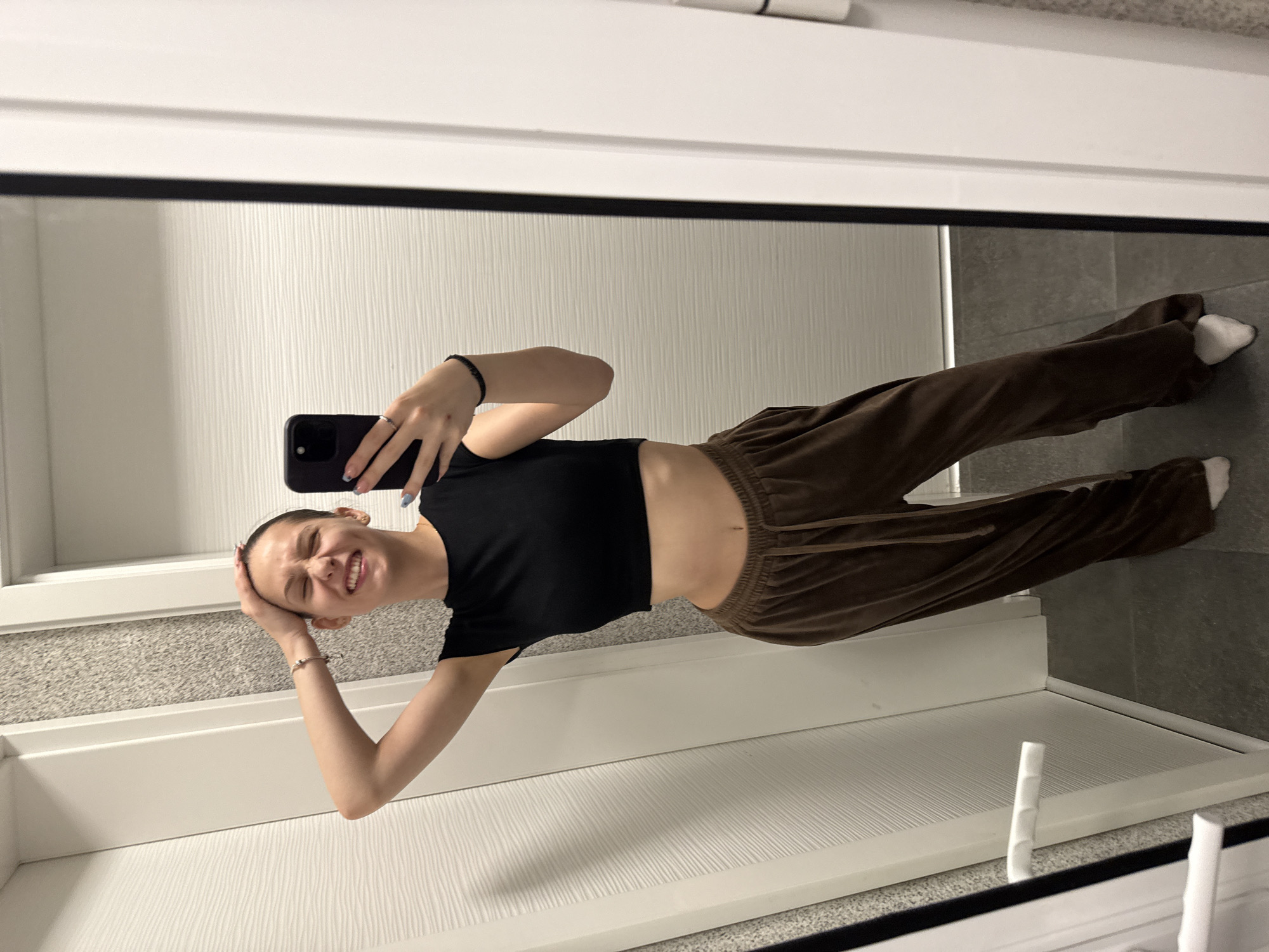A young woman with short hair is taking a selfie in a mirror. She's wearing a black crop top and brown pants. She is positioned within what appears to be a bathroom setting. The environment consists of a white door, white countertop, and gray tiling on the floor and walls. The location appears to be indoors, possibly in a residential building. There is no information to determine if the location is in Sofia, Bulgaria.