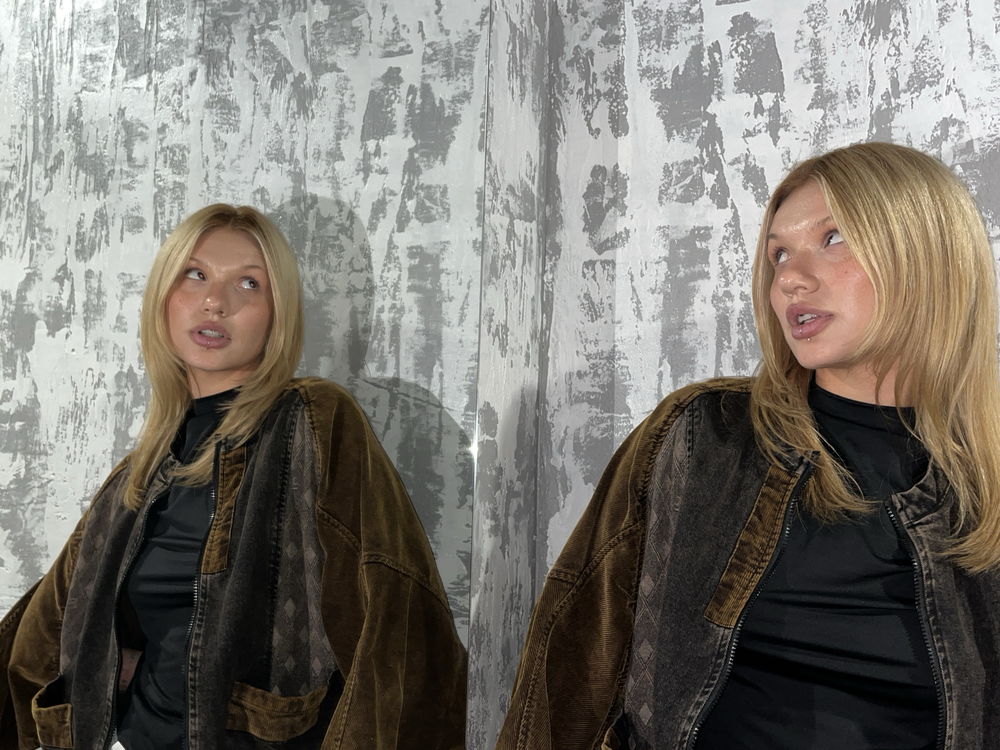 A young woman is depicted indoors, posing against a textured wall with an abstract gray and white paint finish. The right side of the image features a reflective surface, likely a mirror, showcasing her full reflection and creating a symmetrical composition.
The woman has blonde hair, styled with face-framing layers. Both she and her reflection are looking upwards and slightly to their respective sides, with a contemplative or curious expression and mouths slightly ajar. A noticeable lip piercing is visible on her lower lip. She is wearing a dark, possibly black, high-necked top beneath a distinctive jacket. The jacket is oversized and features a combination of textures and colors, including rich brown corduroy on the sleeves and a darker, perhaps distressed, patterned fabric on the body with a subtle diamond motif. It is fastened with a zipper.
The setting appears to be an indoor space with consistent, likely artificial, lighting. There are no external weather cues. The distinctive textured wall serves as the primary backdrop element. No visible text is present in the image. The overall scene suggests a styled photoshoot. The location context provided is Chaikovsky, Russia.