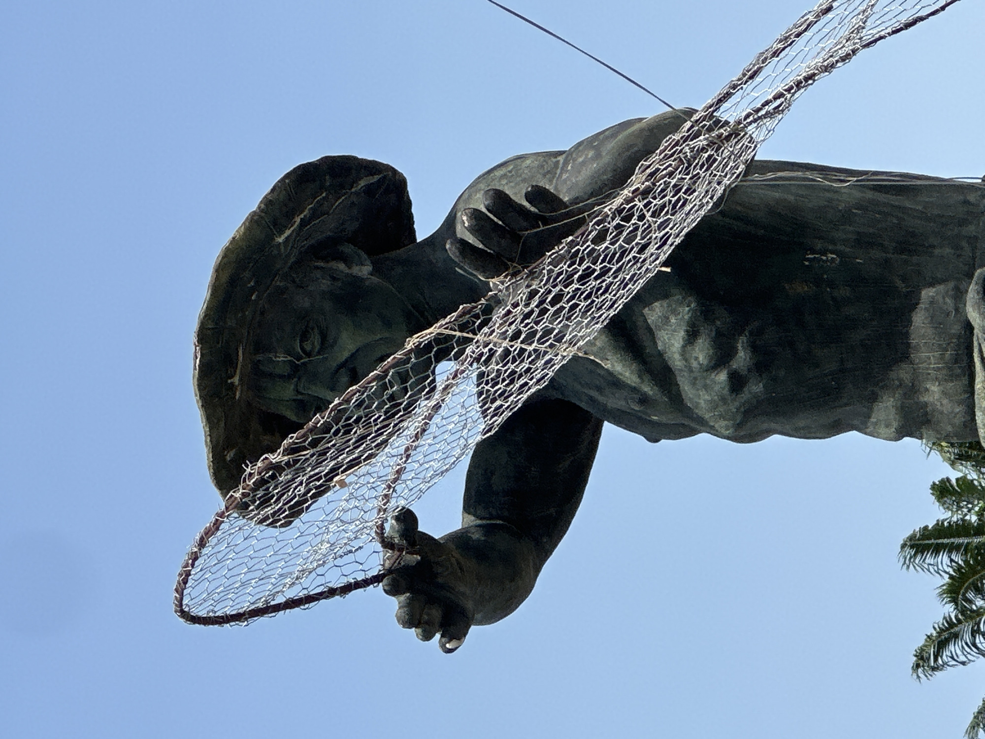 A low-angle shot captures a large, dark-colored statue against a clear, bright blue sky. The statue depicts a human figure, likely male, with a detailed face showing a focused or solemn expression, and appears to wear a hat or head covering. The figure's arms and hands are prominent as it holds a wide fishing net. The net is constructed from a coarse, wire mesh, resembling chicken wire, with a thicker, dark-colored rim. The statue's material suggests weathered bronze or a similar dark metal, with visible texture and contours. The bright, even lighting indicates it is daytime with a sunny sky. In the bottom right corner, a small section of a green palm tree is visible, providing a contextual hint of a tropical or coastal setting. The upward perspective emphasizes the scale and prominence of the statue. Located in Badagry, Nigeria, the statue likely represents a local fisherman or a figure significant to the region's history or maritime culture. No text is visible in the image.