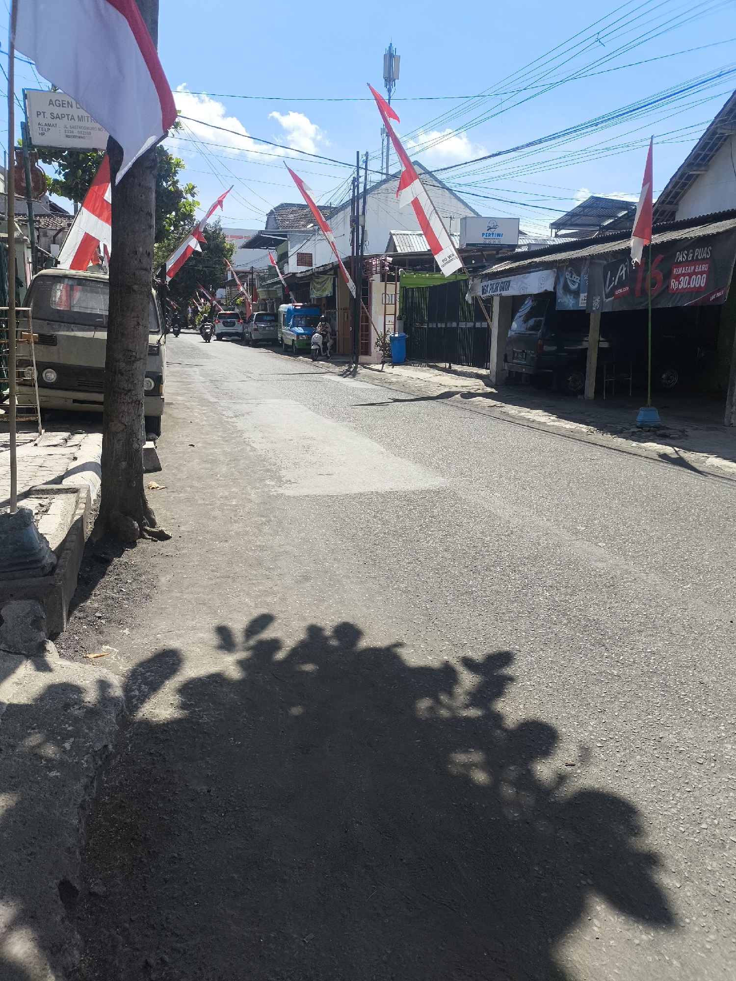 The image depicts a street scene in Citrodiwangsan, Indonesia, under a bright blue sky. Red and white Indonesian flags are prominently displayed on poles, attached to buildings, and strung across the street, suggesting a festive or patriotic occasion. The street is paved and appears to be a busy thoroughfare, with several vehicles visible including a light-colored truck, cars, motorcycles, and a blue van. Buildings line both sides of the street, many with awnings and signs, indicating commercial activity. One sign reads "PT. SAPTA MITRA" and another displays "PAS & PUAS KEMASAN BARU RP30.000". Shadows of trees and leaves are cast across the pavement, indicating the presence of foliage. A tall communication tower is visible in the distance. People are present, with some riding motorcycles and others standing near the buildings. The environment appears to be a mixed residential and commercial area.