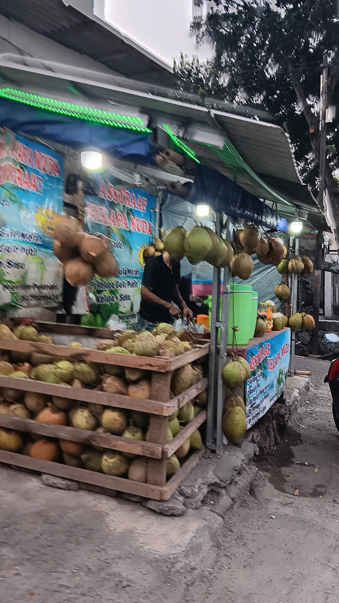 A street-side beverage stall, likely selling fresh coconuts and related drinks, is depicted in South Jakarta, Indonesia. The stall features a wooden structure covered by a corrugated metal roof adorned with green LED strip lights. A blue tarp serves as a backdrop, displaying signage in Indonesian. Piles of green coconuts are prominently displayed, some stacked in wooden crates and others hanging from the stall's canopy. A man in a black t-shirt is visible behind the counter, appearing to prepare a drink. To the right, a large green cooler and a stand with more coconuts are present. The ground is paved with concrete, showing signs of wear and some dampness, suggesting an outdoor urban setting. The overall atmosphere suggests a typical roadside vendor operation.