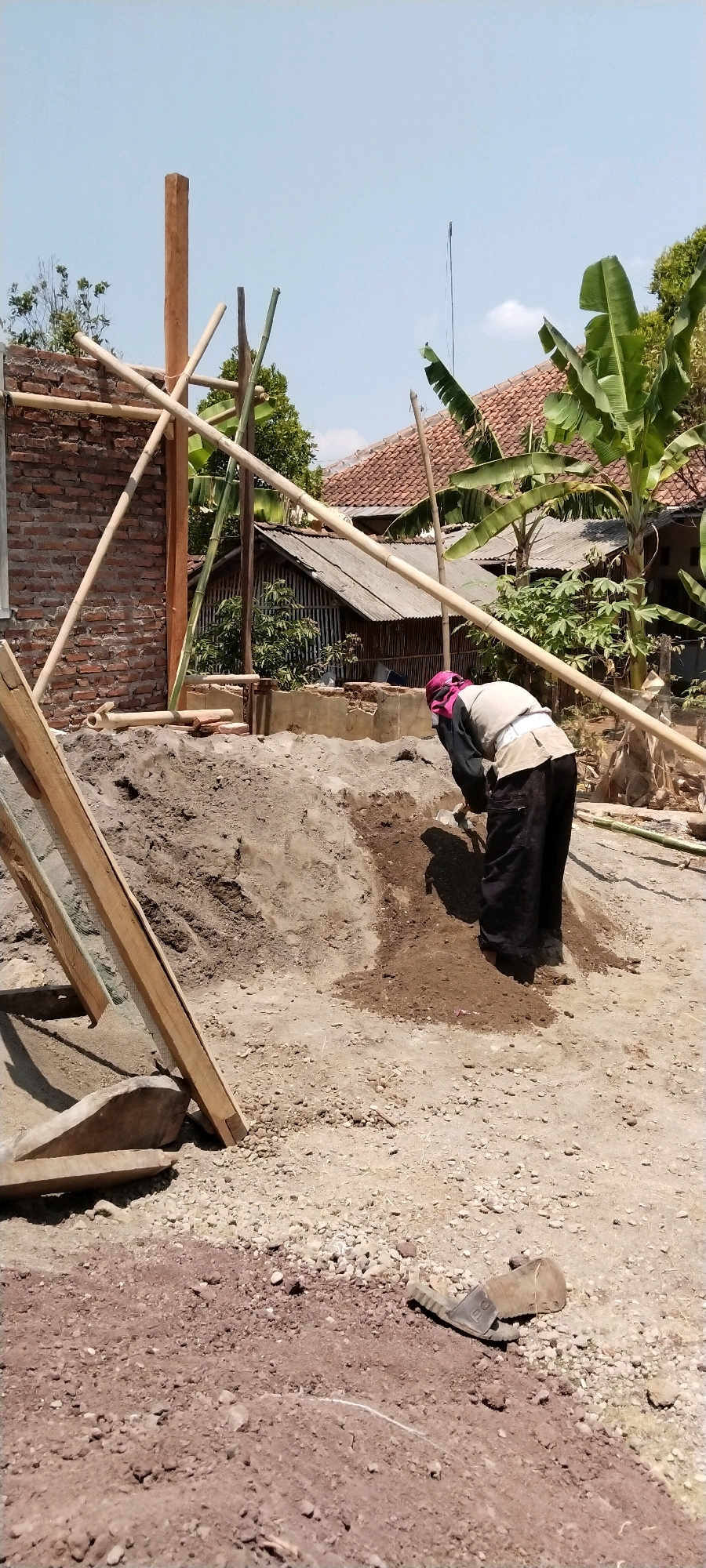In Pamulihan, Indonesia, a construction worker is seen from behind, wearing dark pants and a light-colored shirt, and a pink head covering, as they dig in a pile of dirt. The construction site is evident, with bamboo scaffolding and brick walls visible. In the background, there are residential structures with tiled roofs and banana trees. The ground is a mix of dirt, gravel, and sand. A single sandal lies on the ground to the right of the frame.