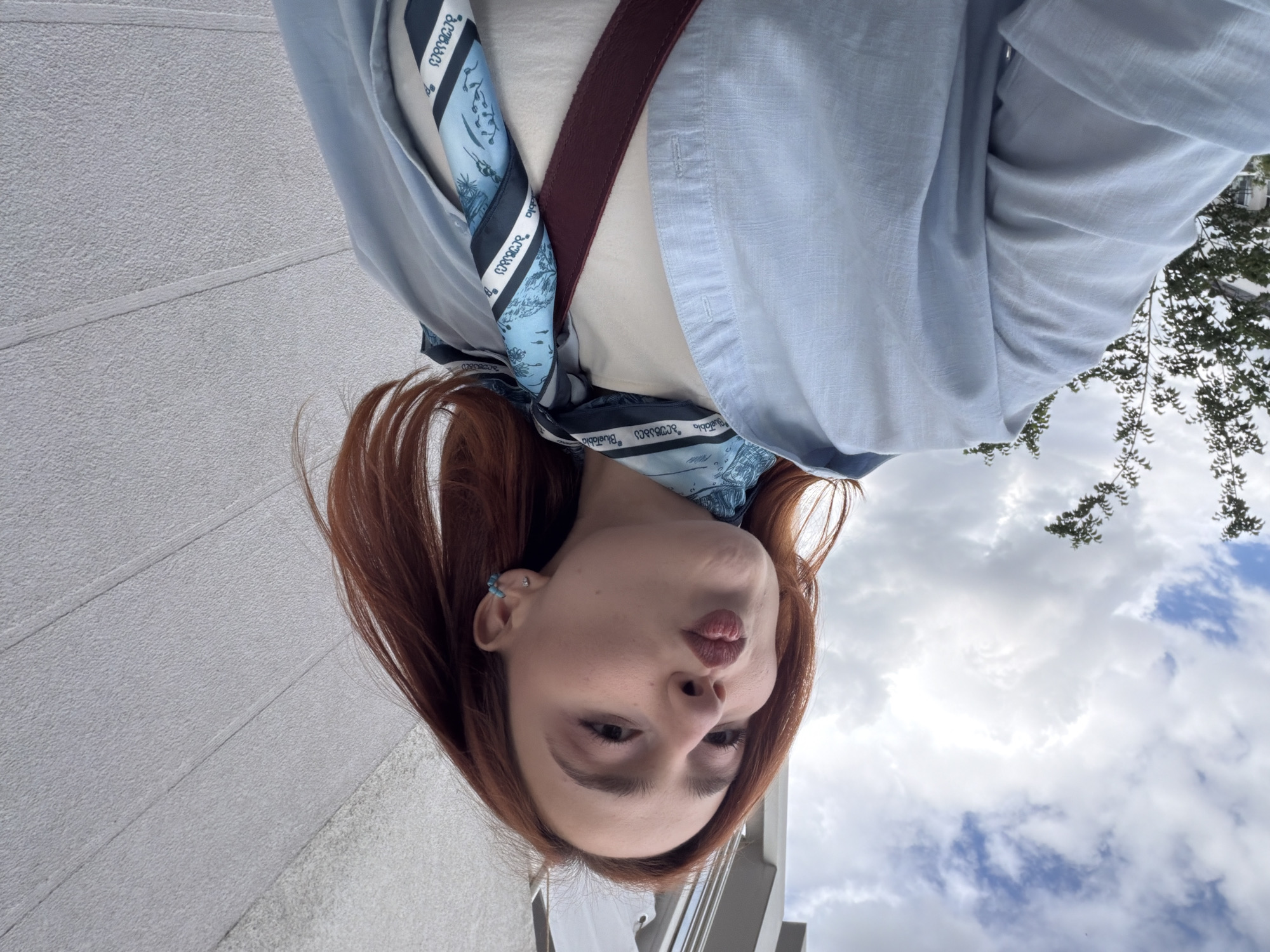 A young woman with reddish-brown hair is depicted in a medium-shot, outdoor portrait on a street in Strasbourg, France. She faces left, offering a pouting expression, and wears subtle eye makeup. Her attire includes a light blue, long-sleeved shirt unbuttoned over a cream-colored top. A blue, white, and dark blue patterned silk scarf, bearing the 'BlueTabia' brand, is tied around her neck. A dark red strap, likely from a bag, crosses her chest. Her visible right ear features a small turquoise hoop earring in the helix and a silver stud in the lobe. The background displays a cloudy sky with patches of blue above the upper left. A light-colored, modern building's top is visible in the upper left-center, partially obscured by green tree foliage. The right side of the image features a light gray, textured wall with horizontal lines.