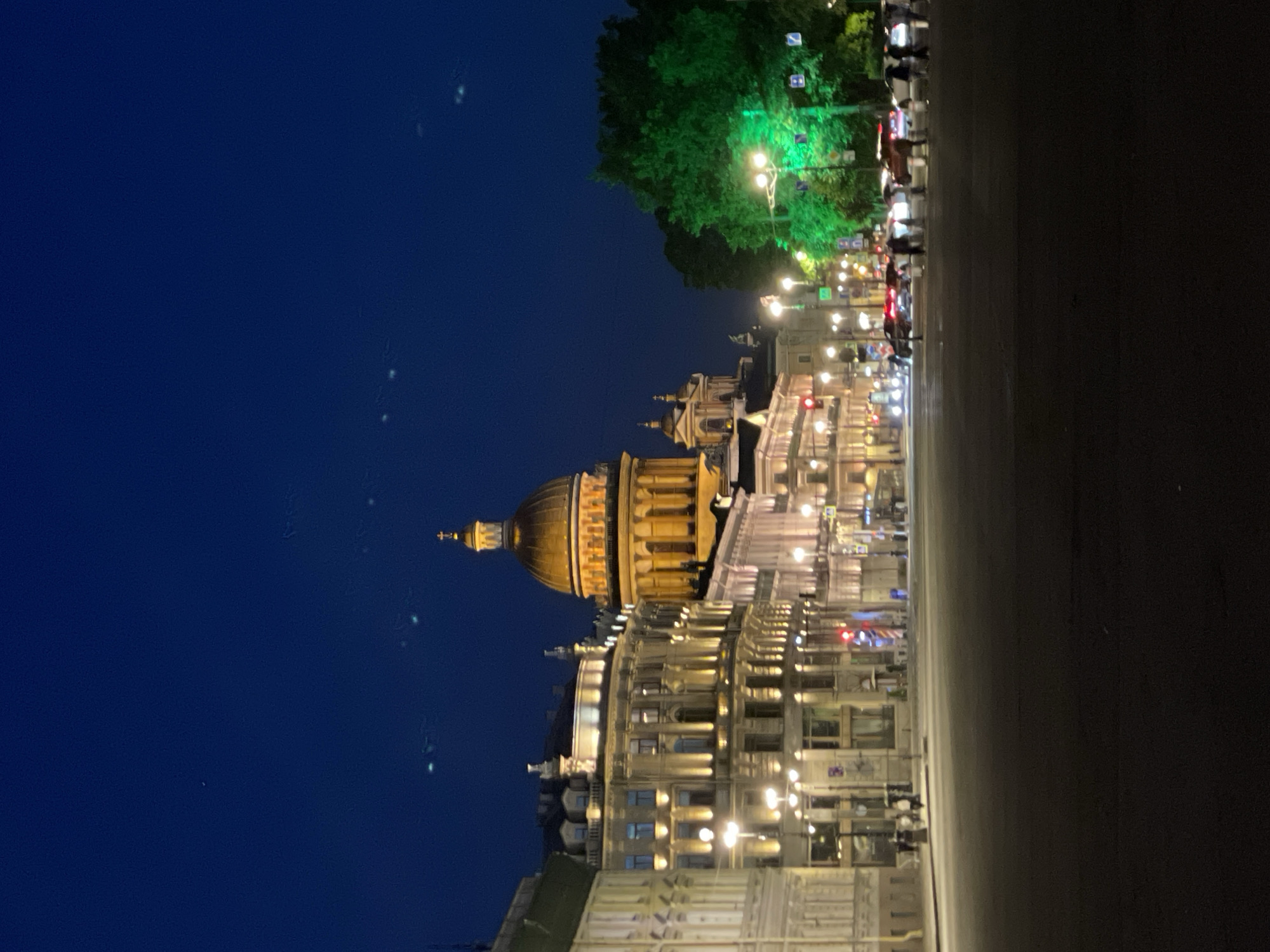 The image depicts a grand urban scene at night or late twilight, characterized by impressive architecture and extensive artificial lighting. A prominent building with a large golden dome and classical columns stands on the left, brightly illuminated against the deep blue sky. Adjacent to it, a series of multi-story buildings line what appears to be a wide street or plaza, their facades detailed with ornate architecture and numerous glowing windows.
To the upper right, a large, dense green tree is brightly lit by multiple streetlights, which also cast a warm glow over the entire scene. A broad, dark street occupies the foreground and extends towards the right, with several red traffic lights visible in the distance. Blurry shapes in the background suggest the presence of people or vehicles, indicating ongoing urban activity. Poles with indistinct signs are also visible along the street. The overall atmosphere is one of a majestic city, likely European due to the architectural style, beautifully highlighted by the night lights. No discernible text is visible.