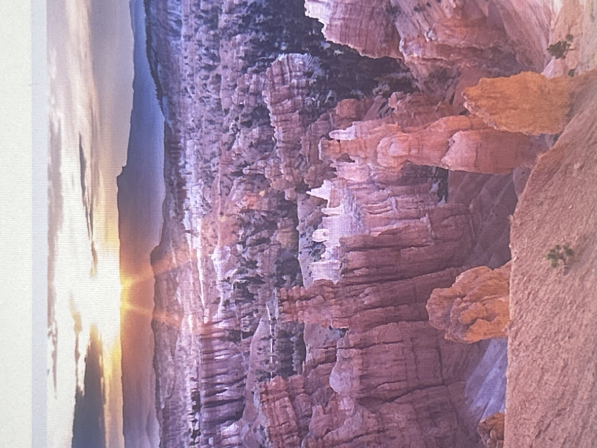 The image depicts a panoramic view of Bryce Canyon National Park in Utah, USA, at sunrise. The foreground is dominated by the vibrant orange and pink hues of Hoodoos, tall, slender rock spires formed by erosion. These formations are densely packed, creating a surreal, otherworldly landscape. In the mid-ground, the canyon floor is visible, dotted with pine trees. The background features layers of distant mountains and mesas, bathed in the soft, golden light of the rising sun. The sky above is a gradient of pale blue and soft gray, with streaks of clouds catching the sun's rays. The overall impression is one of natural grandeur and serene beauty. There are no people or man-made structures visible in the image. The location is explicitly identified as Bryce Canyon National Park, Utah, USA, and not Sofia, Bulgaria.
