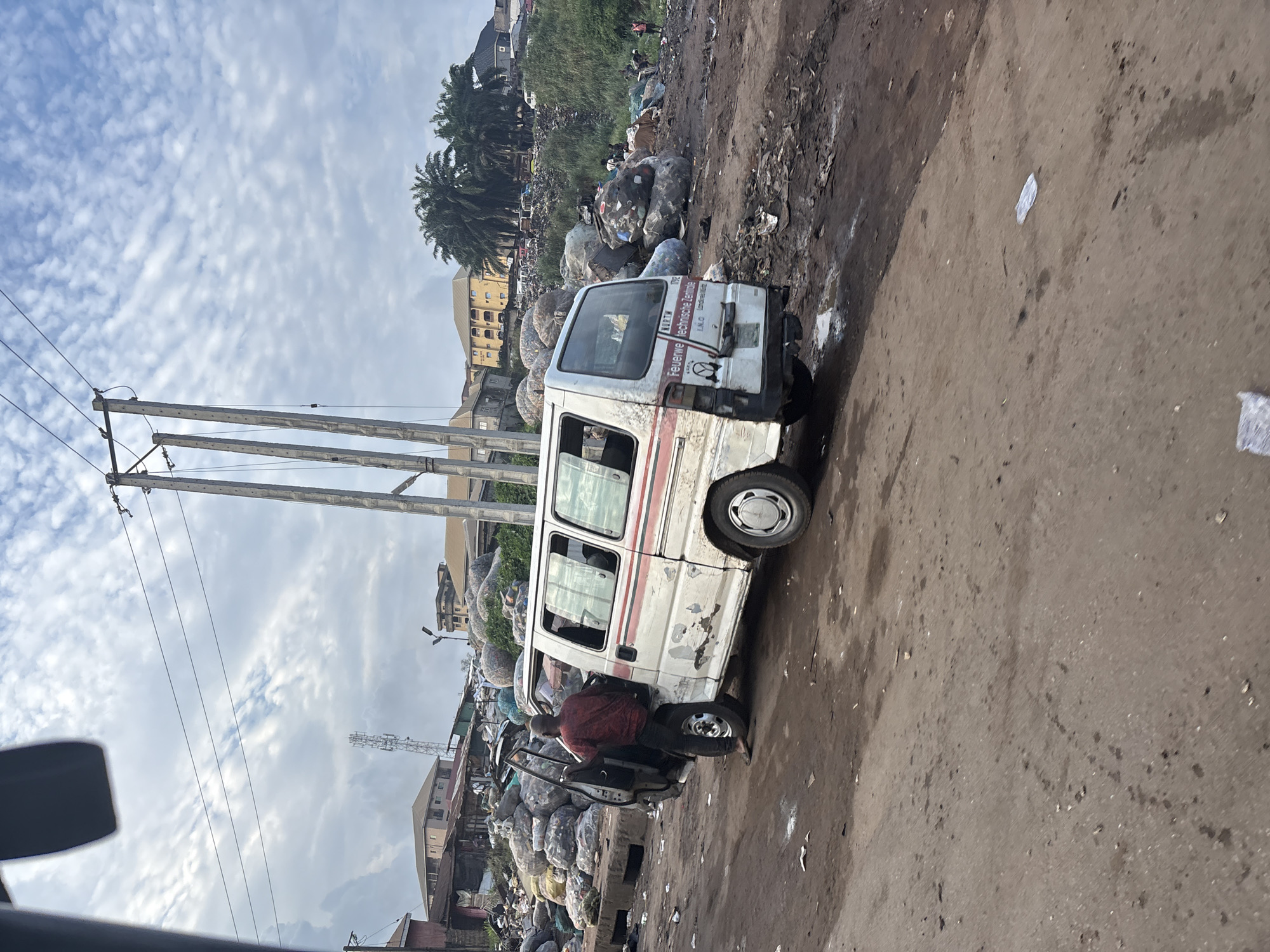 In Onitsha, Nigeria, a white van with red and black trim is parked on a dirt road. The van is weathered and shows signs of damage. A man in a red shirt and dark pants stands with the driver's side door open, appearing to be interacting with the van. The background is dominated by large piles of what appears to be refuse or compressed materials, possibly plastic bales, creating a landscape of waste. Buildings and trees are visible in the distance, suggesting an urban or semi-urban environment. Electrical poles and wires are present in the sky. The ground is uneven and covered in dirt and debris, indicating a less developed or potentially neglected area.