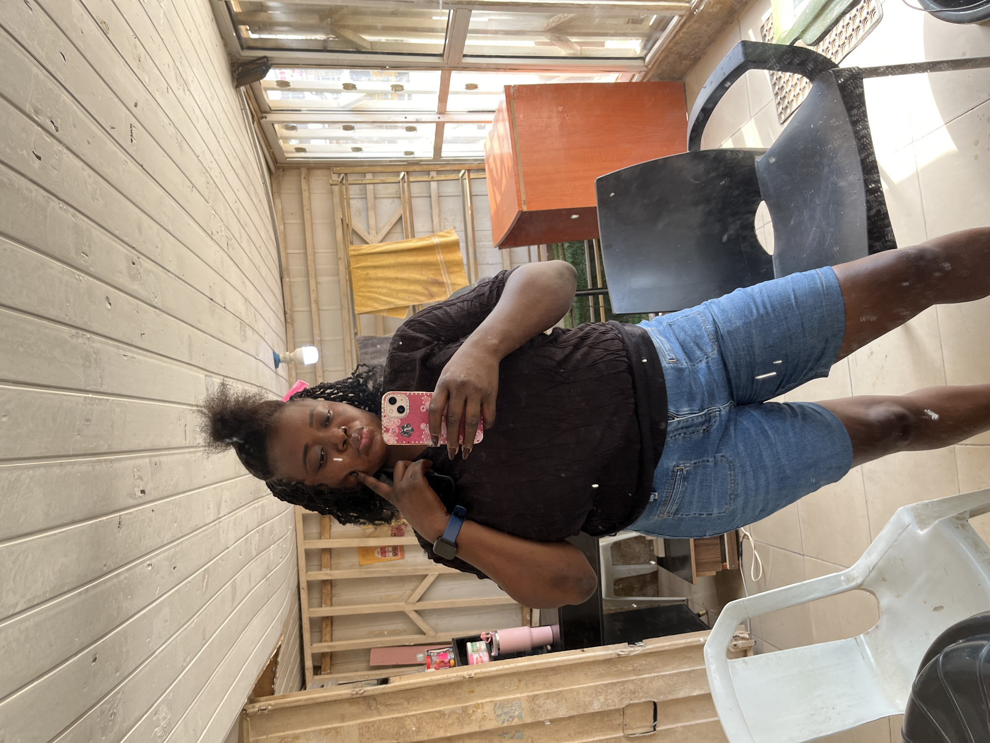 A woman is taking a selfie in what appears to be a sunroom or conservatory undergoing renovation. She is positioned in the center of the frame, facing the camera. She has dark, braided hair pulled into a high ponytail with some strands loose around her face. She is wearing a dark, textured long-sleeved top and light blue denim shorts. She is holding a pink phone with a floral pattern in her right hand, which is angled to capture her reflection in a mirror. Her left hand is resting on her cheek, and she has a slight smile. A blue smartwatch is visible on her left wrist.
The background shows a construction or renovation environment. There are unfinished wooden beams and panels on the walls and ceiling. Some yellow sheeting is visible on a wooden frame in the background. To the right of the woman, there is a large, dark plastic chair. Further back, an orange box-like structure is visible, along with other construction materials and debris. The floor is tiled with light-colored tiles. The presence of a skylight or glass ceiling suggests it is daytime. The overall impression is one of a casual, possibly domestic, setting in the midst of improvement work.
There are no visible text elements in the image. The lighting suggests it is daytime, likely with sunlight filtering through the glass ceiling. The location context of Abuja, Nigeria, is provided but not visually evident within the image itself.