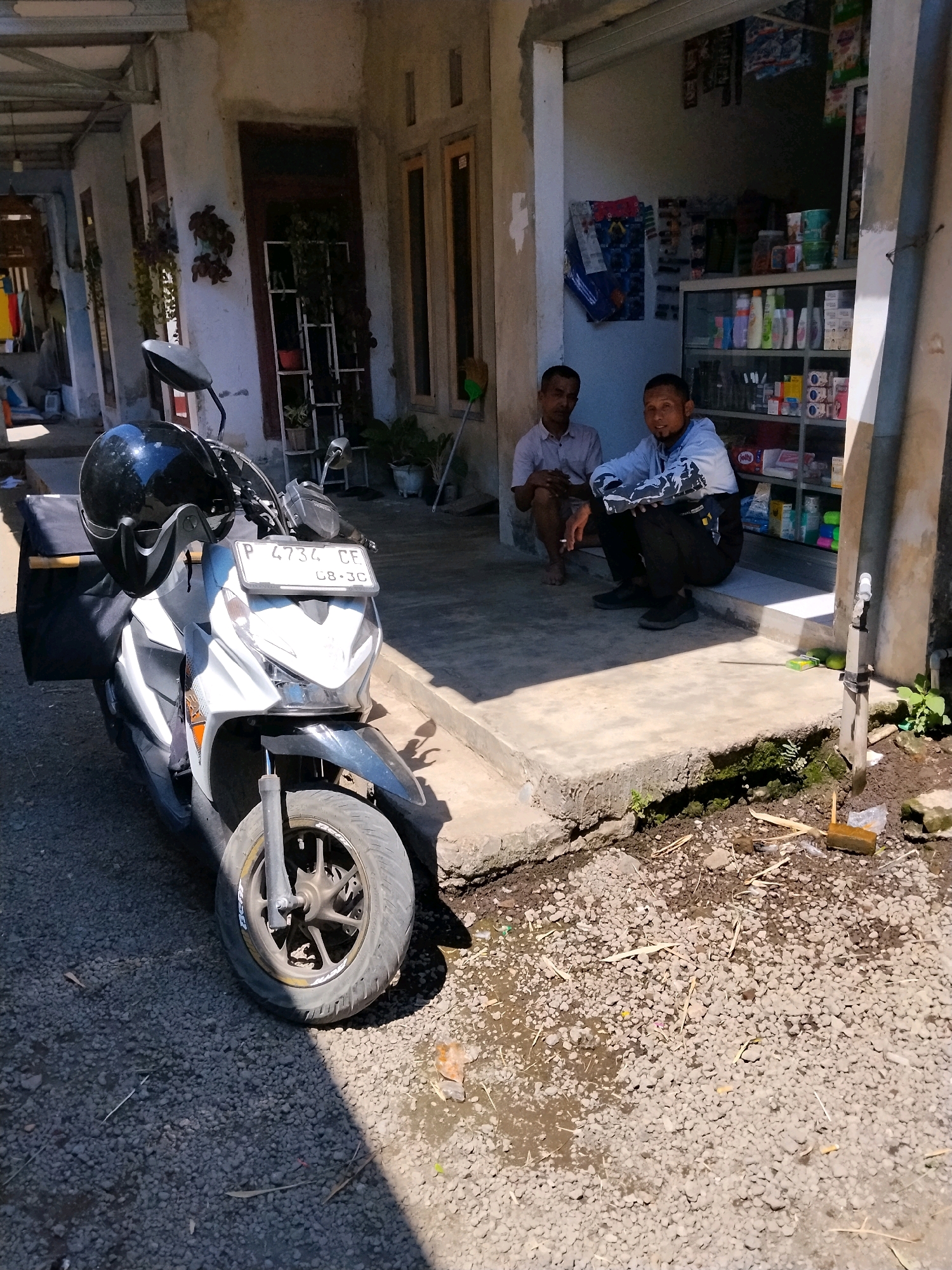 In Panggungan, Indonesia, a white and gray scooter with a black helmet secured to the handlebars is parked on a gravelly surface in front of a small storefront. Two men are seated on the step of the store. The man on the left, wearing a light-colored collared shirt, is partially obscured and appears to be smoking. The man on the right, wearing a light blue hooded jacket with a camouflage pattern on the sleeves and black pants, is looking towards the camera. Behind him, the store is stocked with various items on shelves, suggesting it is a small convenience store or pharmacy. The building features a white facade with wooden-framed windows and doorways. The environment is outdoors, with sunlight casting shadows on the ground.
