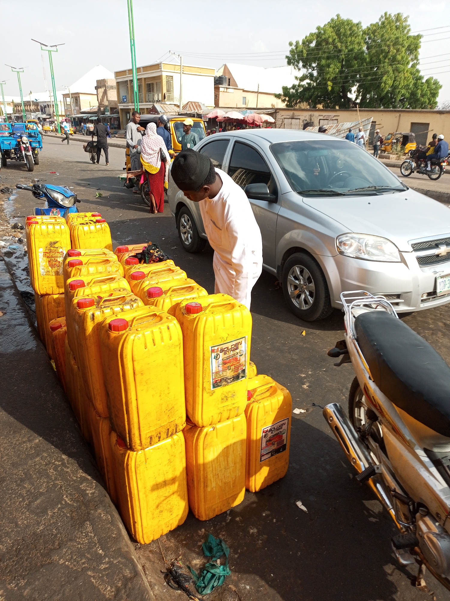 The image shows an outdoor scene, possibly a street in a busy market or urban area. In the foreground, there is a stack of yellow jerrycans, possibly containing a liquid substance like gasoline or cooking oil, arranged in multiple rows. To the right of the jerrycans, there's a silver-colored car, with a man in a white robe and a black hat standing near the front of the car. A motorcycle is parked next to the car. In the background, there are people walking and riding various vehicles, including auto rickshaws (tuk-tuks). Buildings with simple architectural designs are visible, and there are overhead light poles lining the street. The overall impression is a typical street scene in a developing urban area, with commercial activity and pedestrian/vehicle traffic.