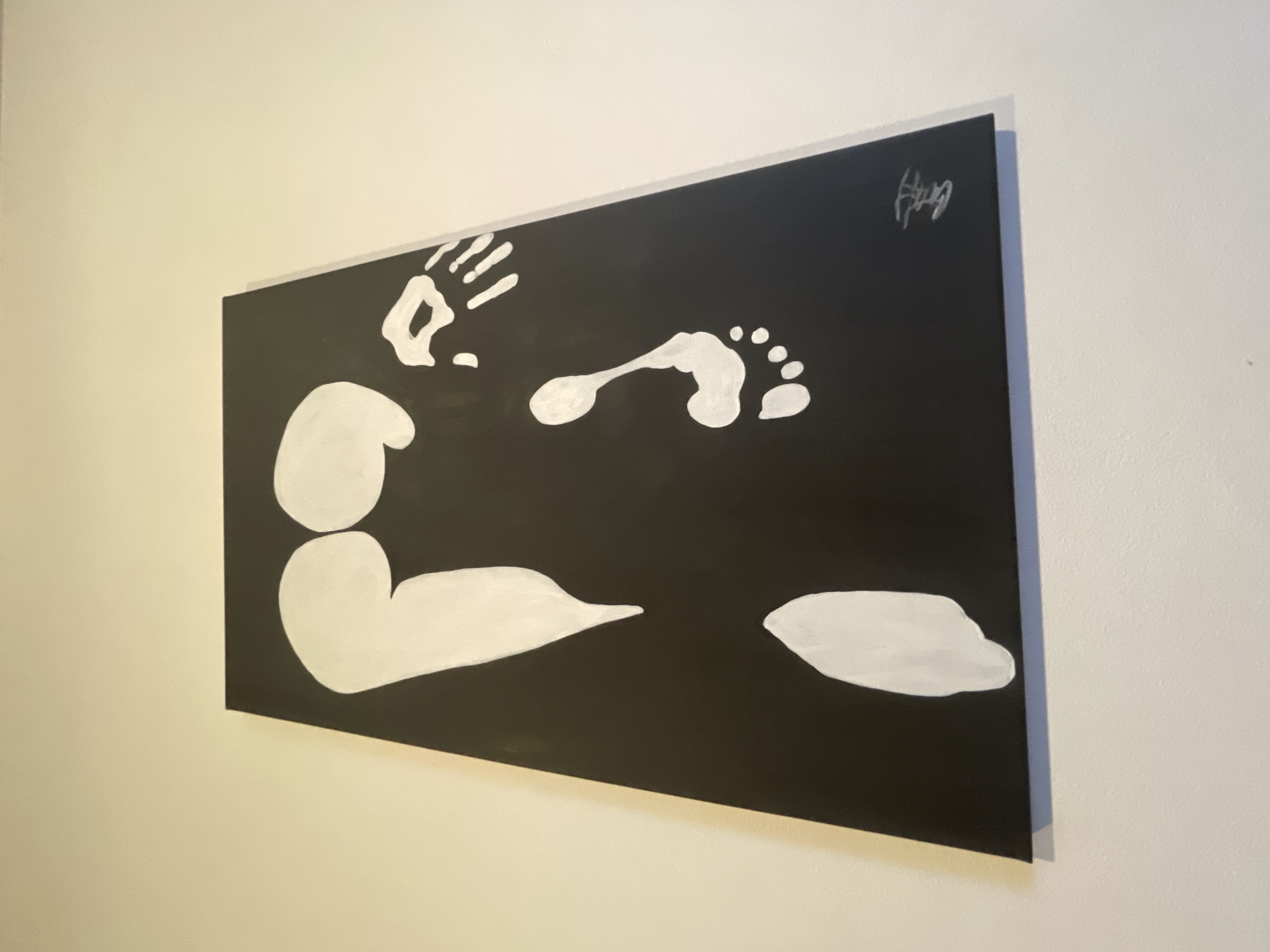 A black and white painting is displayed on a pale wall. The painting features a white handprint, a footprint, and abstract shapes suggesting a head and body. A signature is visible in the upper right corner. The painting is mounted on a wall, likely indoors. Location information is not available from the image.