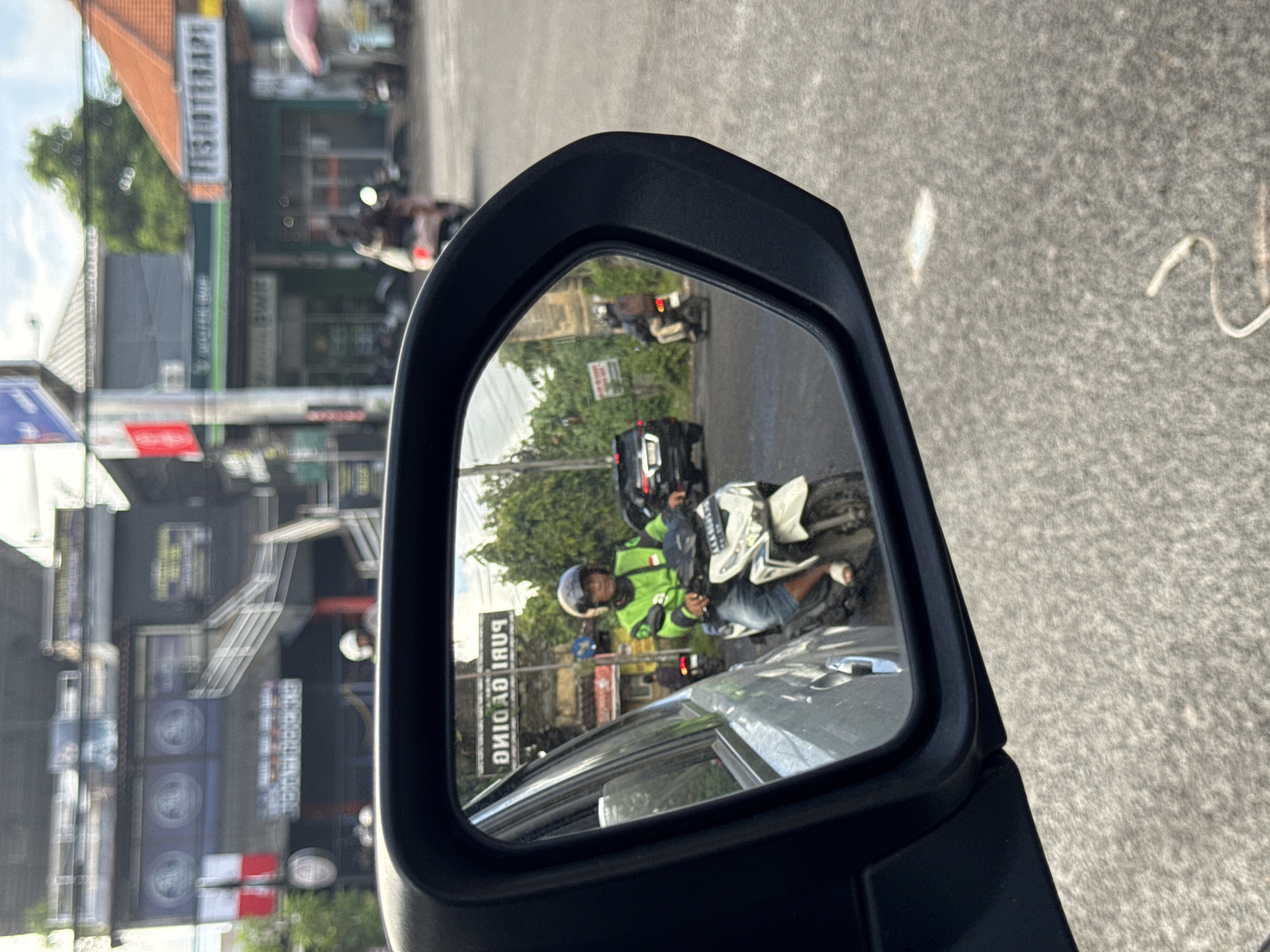 The image is a view from a car side mirror in Kuta Selatan, Indonesia. The mirror shows a motorcyclist in a green jacket and helmet on a white motorbike, driving on the road behind the car. A black vehicle is partially visible behind the motorcyclist. In the background, blurred buildings and signs are present, including a sign that reads "PURI GADING". The road visible in the mirror appears to be paved. The mirror itself is black and has a sharp, angular design. The exterior part of the image reveals a grey asphalt road and a blurred cityscape.