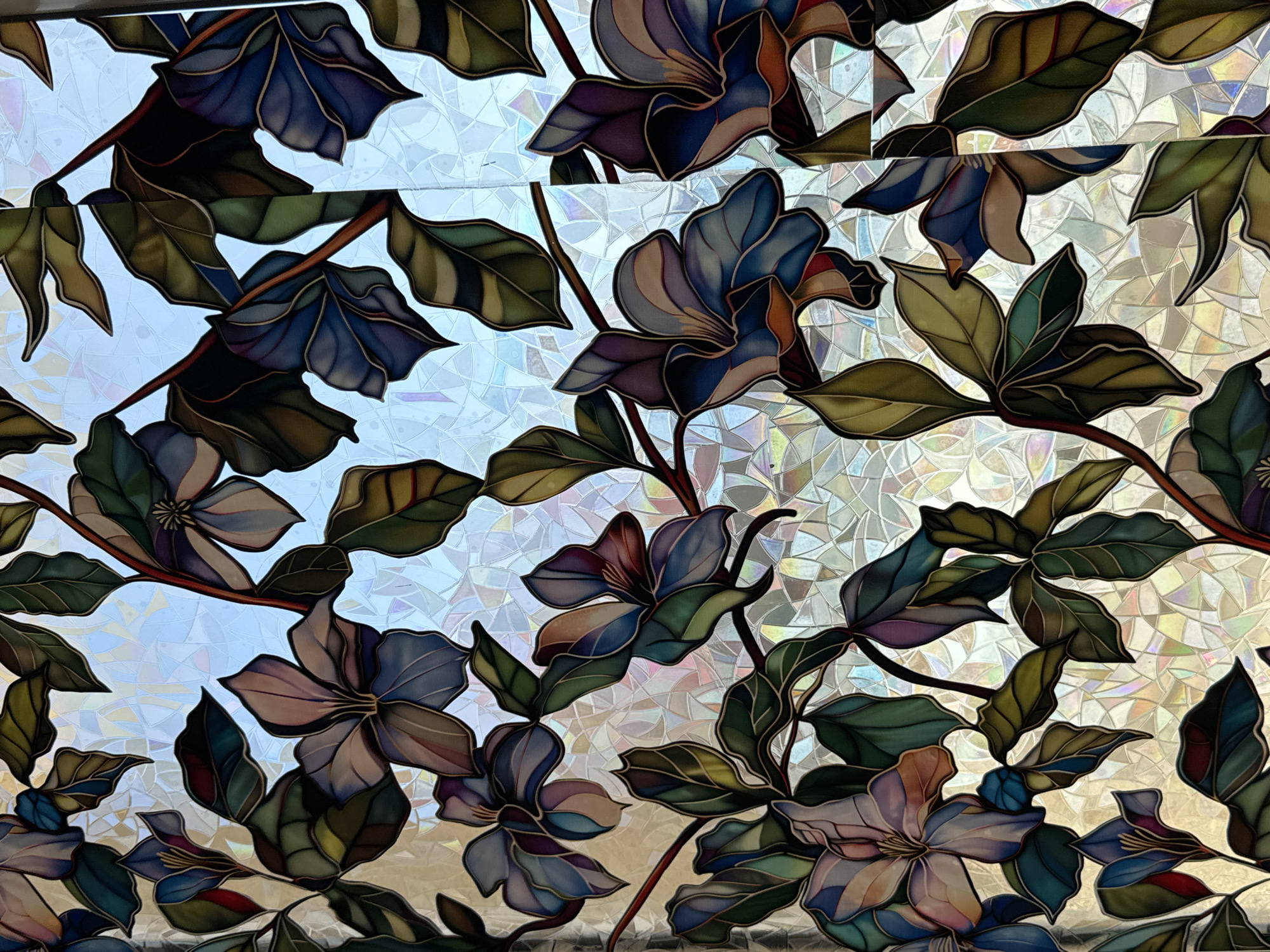 This media file showcases a close-up view of stained glass featuring a floral design. The subjects are stylized flowers and leaves rendered in a variety of colors including deep blues, purples, greens, and earthy browns, with highlights of lighter blues and pinks. The floral elements are depicted on dark, thin stems, intertwined against a background of textured, translucent glass that diffuses light with an iridescent, crystalline pattern.
The stained glass appears to be a decorative panel, possibly a window or a partition, given its translucence and the way light passes through it. There are no people or animals visible. The scene is static, depicting a naturalistic yet artistic interpretation of flowering branches. The colors suggest a rich and vibrant aesthetic.
Notable details include the intricate detailing within the petals and leaves, giving them a sense of depth and form. The background glass, with its tessellated pattern of small, irregular shapes, creates a shimmering effect. The overall impression is one of intricate craftsmanship and artistic beauty, evoking a sense of calm and perhaps a touch of opulence. There is no visible text within the media file. The time of day and weather are not discernible from the image, as the light source appears to be internal or uniformly diffused through the stained glass itself. The location is also unknown, but the style might suggest an indoor setting such as a home, a place of worship, or a commercial establishment where decorative glass is used.
