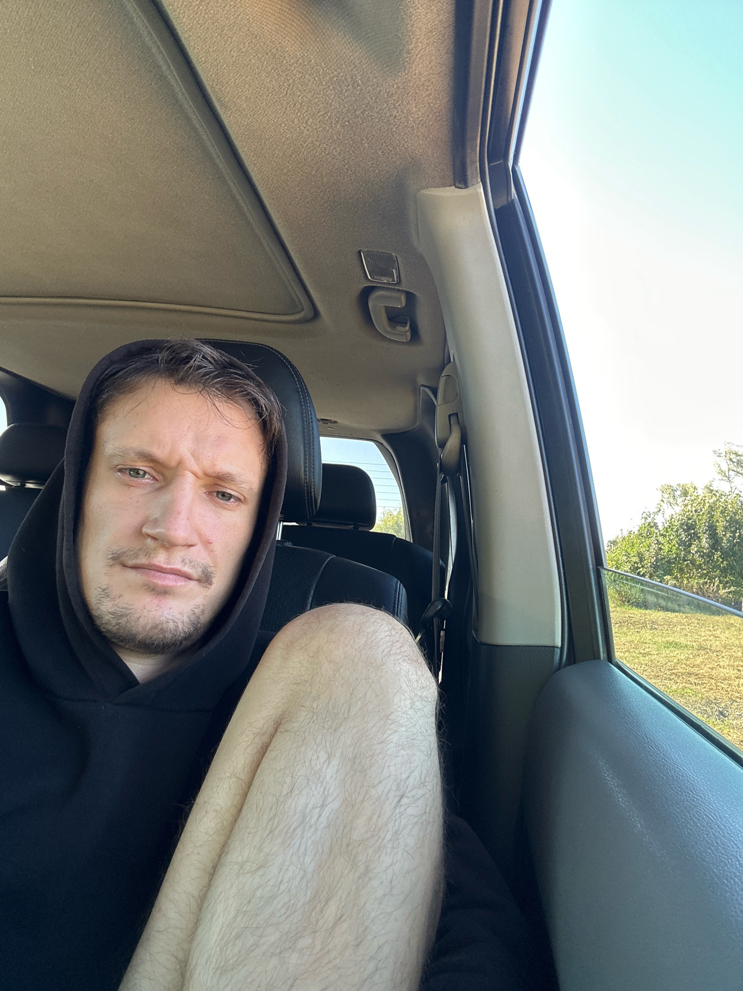 A man wearing a black hooded sweatshirt sits in the passenger seat of a vehicle, looking towards the camera with a neutral expression. His right leg is visible, bent and positioned in the foreground. The interior of the car, including the sunroof, headliner, and seatbacks, is visible. Outside the open window, a grassy field and trees are illuminated by sunlight. The image is a selfie, likely taken while traveling. The scene is not set in Makiv, Ukraine.