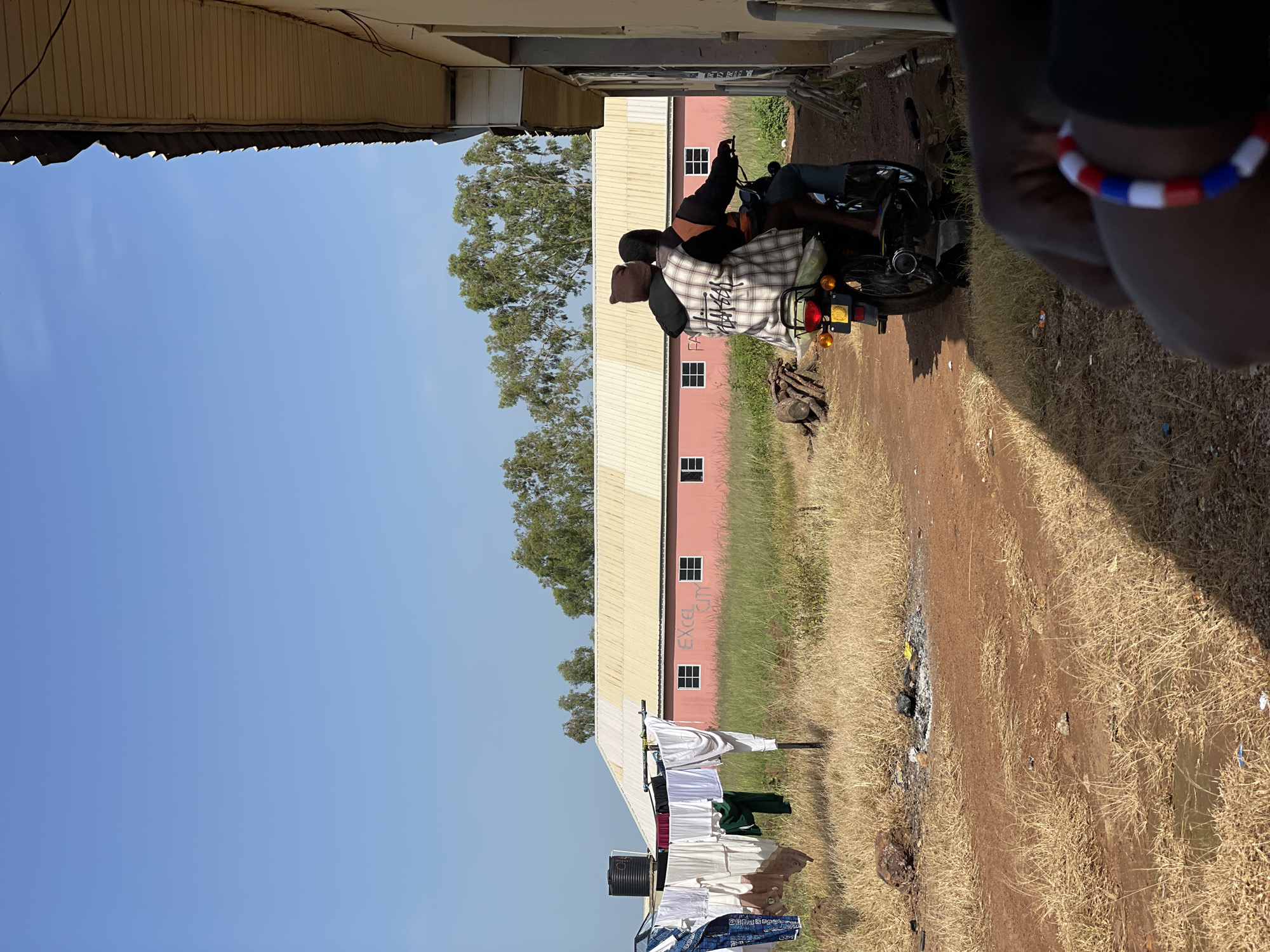 In the city of Durumi, Nigeria, a clear blue sky stretches over a scene dominated by dry, golden grass and a dirt path. To the left, a clothesline hangs with various garments, including blue patterned fabric, white shirts, and a dark green item, adjacent to a black water tank. A pink building with a white-patterned roof and multiple evenly spaced windows is visible in the background, with the words "EXCEL CITY" spray-painted in red on its facade. Trees with green foliage are scattered behind the pink structure. In the mid-ground, two individuals are riding a motorcycle. The rider wears a dark hat and an orange garment, while the passenger is dressed in a plaid shirt and a brown knitted hat. They are moving away from the viewer. In the foreground, to the right, a portion of a building with a light yellow exterior and a weathered, dark roof is visible, casting a shadow onto the ground. A person's arm, adorned with a red, white, and blue beaded bracelet, is also in the foreground, suggesting they are the photographer or an observer. The ground is rough, with patches of dry grass, scattered rocks, and evidence of a small fire pit with ash and debris.
