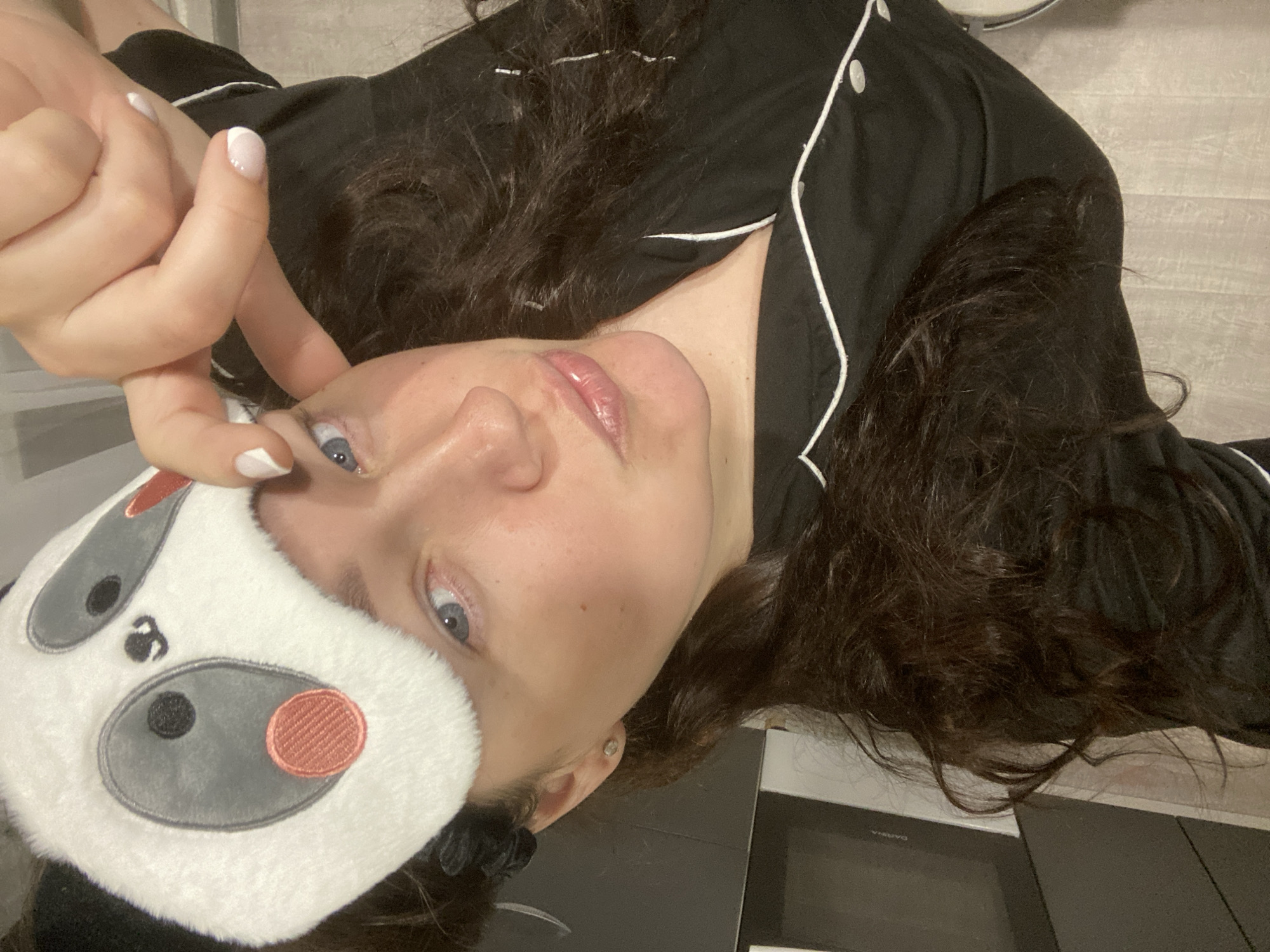 A young woman lies on a surface, possibly a bed or countertop, wearing black pajamas with white piping and a white plush eye mask shaped like a panda bear. Her dark, curly hair is spread around her head. She holds the eye mask partially over her face with her left hand, which has white-tipped nails. She is looking directly at the camera with light-colored eyes. The background is blurry and appears to be a light-colored wall. There is a dark surface in the lower right corner of the image, potentially part of a countertop or appliance. The location of this image is not identifiable to the city of Sochi, Russia.