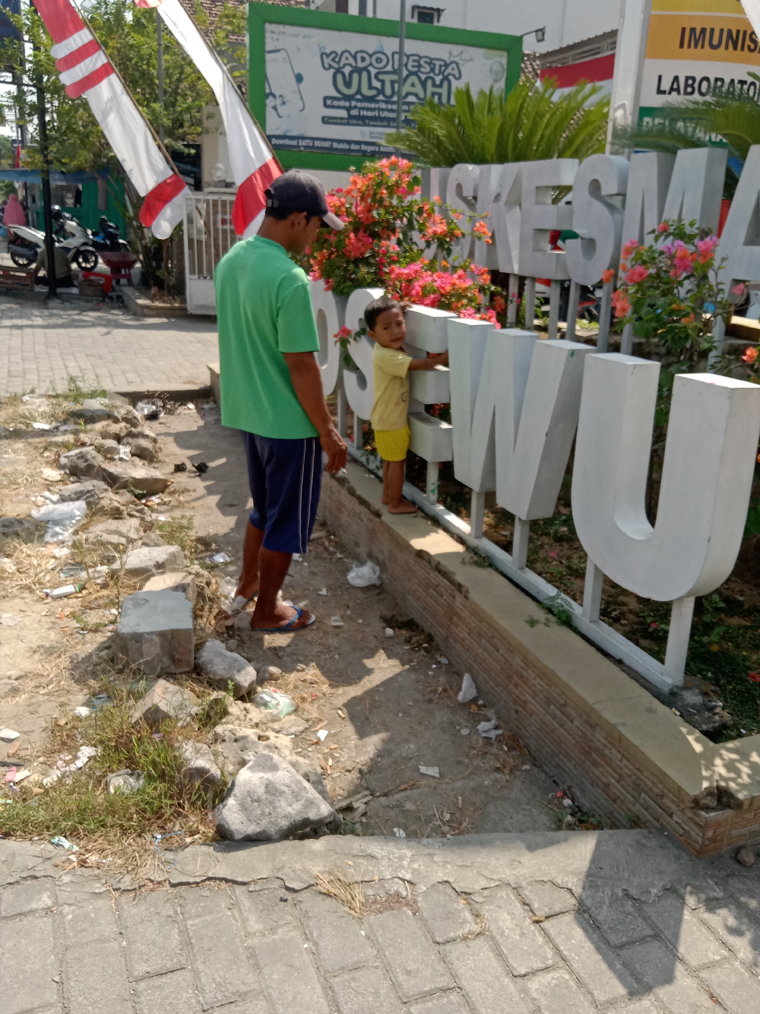 In Klepek, Indonesia, a man in a green t-shirt and blue shorts stands on a dusty path next to a large white three-dimensional sign that reads "PUSKESMA". A young child in a yellow t-shirt and shorts is positioned on the other side of the sign, leaning against it. Behind the sign, a building with a green facade is visible, displaying text that includes "KAPORESTA ULTAH" and "IMUNISASI LABORATOR". Red and white flags flutter in the background. The ground around the sign is littered with debris, including rocks and trash. The scene appears to be outdoors on a sunny day, with shadows cast by the objects and people.