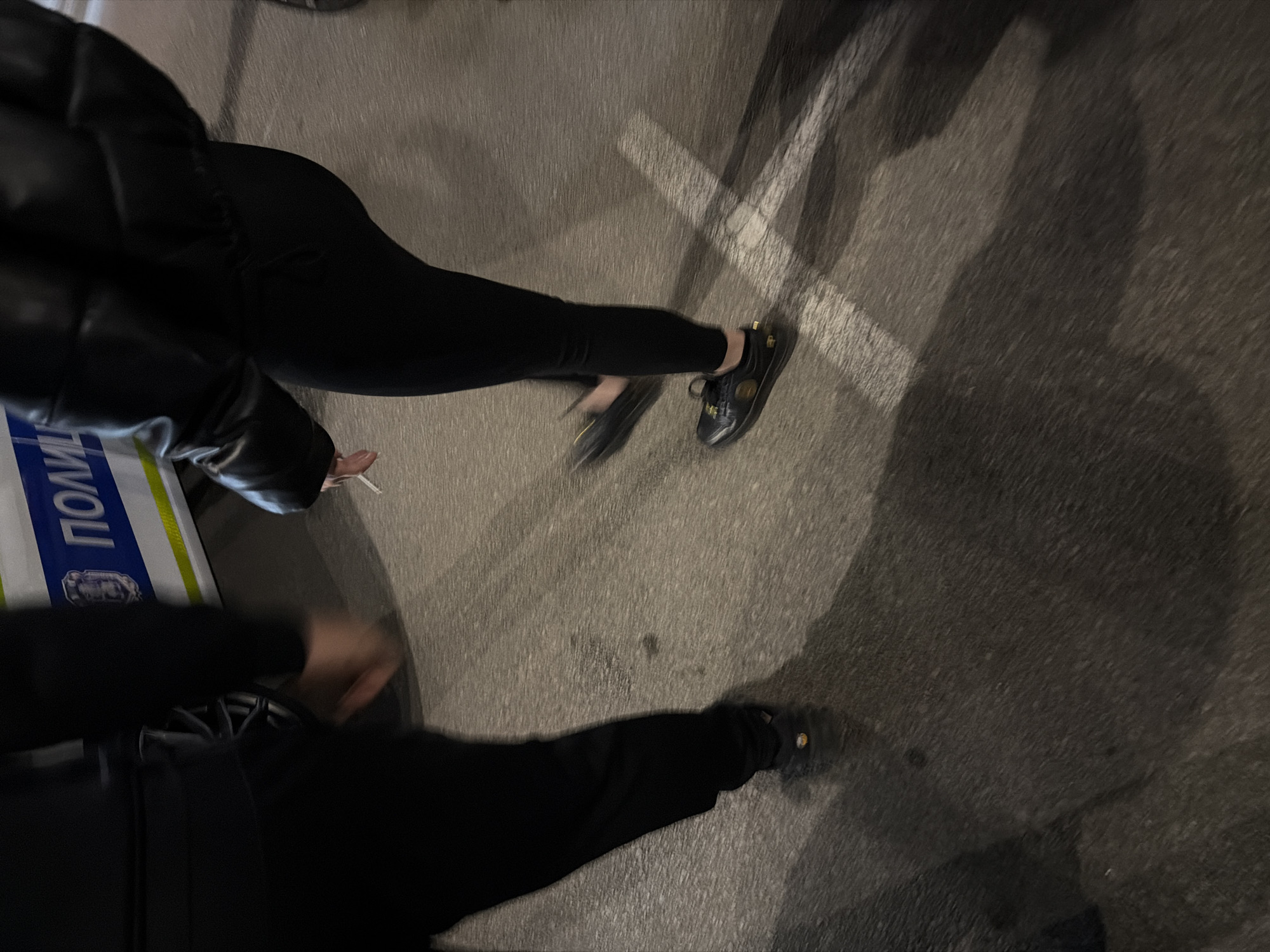 The image shows an aerial view of two individuals standing on an asphalt surface, potentially a street, in Sofia, Bulgaria, as indicated by the presence of a police vehicle with the Cyrillic inscription "ПОЛИЦИЯ" (Police). The upper figure wears a black puffer jacket and black leggings, with black shoes. The individual holds a lit cigarette. The other person is partially visible at the bottom, wearing black pants and shoes. White lines are painted on the asphalt. The photograph appears to be taken at night or in low light conditions.