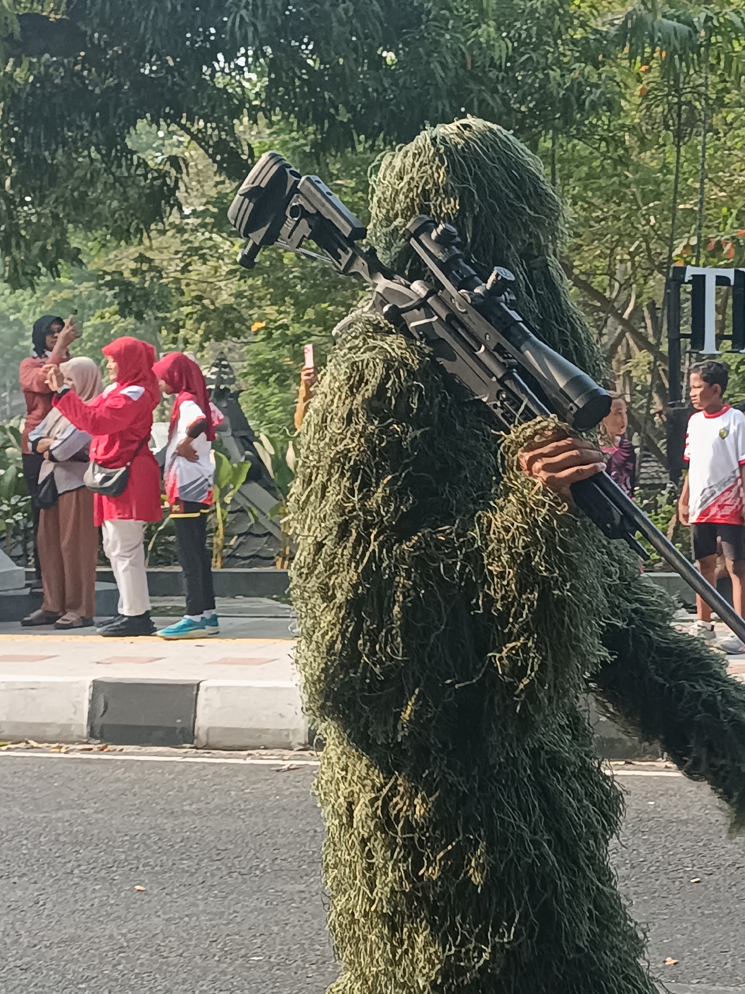 In Sleman, Indonesia, a person in a ghillie suit, designed to resemble foliage, stands with a sniper rifle slung over their right shoulder. The rifle has a prominent scope. The individual is positioned in the foreground, partially obscuring the background.
Behind the person in the ghillie suit, a group of people are visible standing on a sidewalk next to a road. From left to right, the first individual wears a brown headscarf and a brown top with a white pattern. Next to them, a person in a red hijab and red jacket wears white pants and black shoes, holding a mobile phone to take a picture. Following them, a person with a red hijab wears a white and red top and black pants with blue shoes. Further to the right, another person's hand is visible holding a mobile phone. Beyond this group, a young boy in a white shirt with red accents and black shorts stands on the right side of the image.
The environment appears to be outdoors with trees and foliage providing a green backdrop. Part of a white and black striped barrier is visible along the edge of the sidewalk, and a paved road is in the lower portion of the frame. The scene is illuminated by natural daylight, casting shadows and suggesting it is daytime. There is also a partial view of a sign with a large "T" on it in the background.