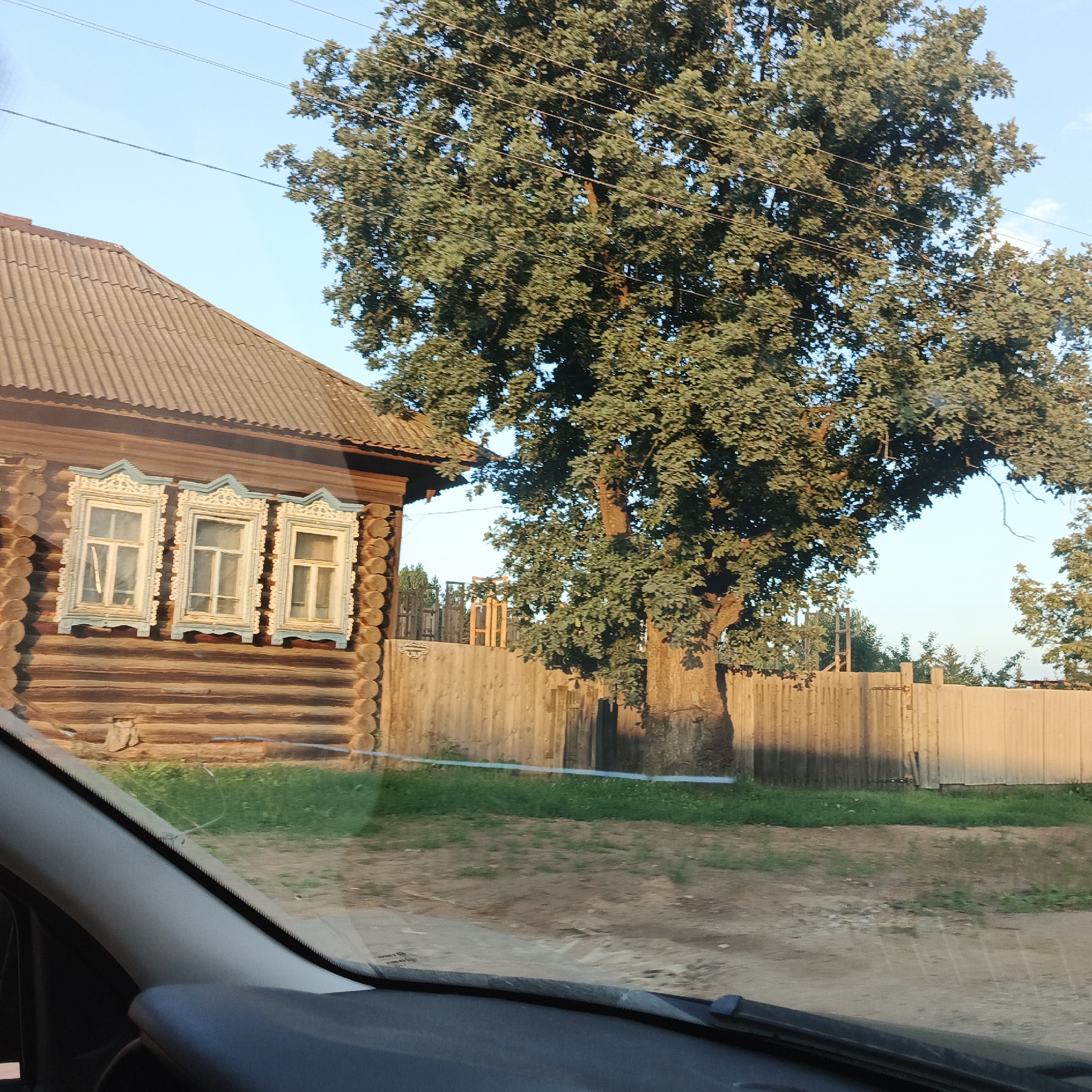 The image depicts a rural scene in Vanki, Russia, featuring a traditional log house with intricately carved window surrounds. The house has a corrugated metal roof. Adjacent to the house stands a large, mature tree with lush green foliage. A wooden fence runs along the property line, separating it from a dirt road and some sparse grass and vegetation. The sky is clear and blue. The foreground shows a portion of a car's interior, including the dashboard and windshield wiper, suggesting the photo was taken from inside a vehicle. The overall impression is of a peaceful, rustic setting.