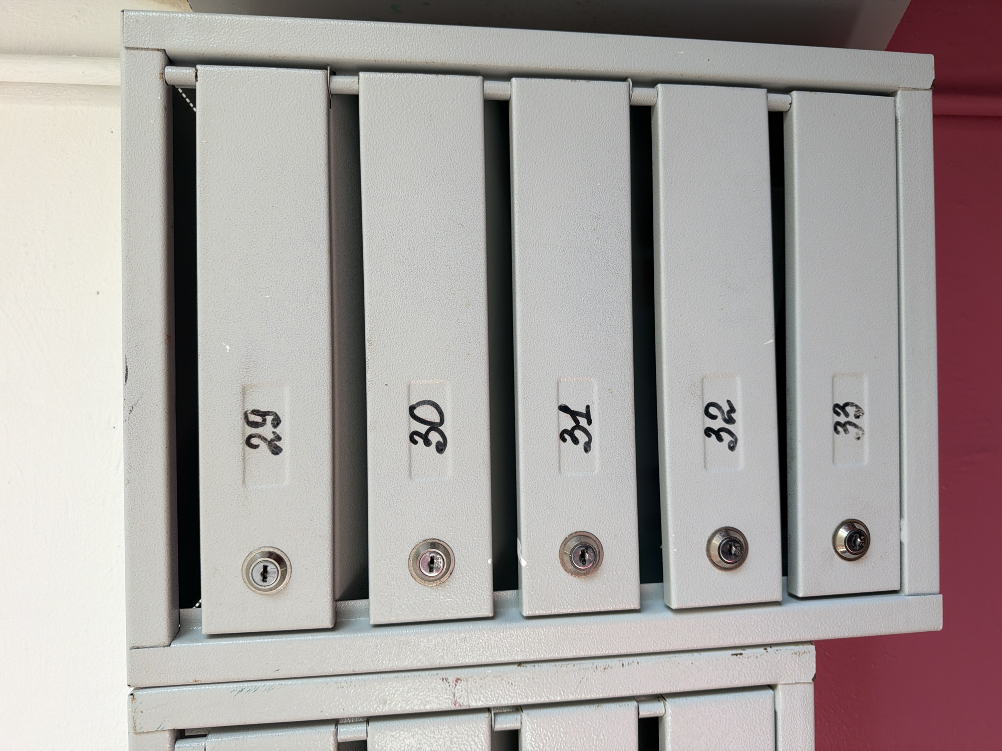 The image shows a bank of light gray metal mailboxes with individual compartments. Each compartment has a small keyhole on the left and a black handwritten number on the right. The visible numbers are 29, 30, 31, 32, and 33. The mailboxes are installed in an interior setting, against a white wall above a pink baseboard. This appears to be a typical scene in an apartment building in the city of Okha, Russia, where such mailboxes are common. There are no people or animals visible in the image.