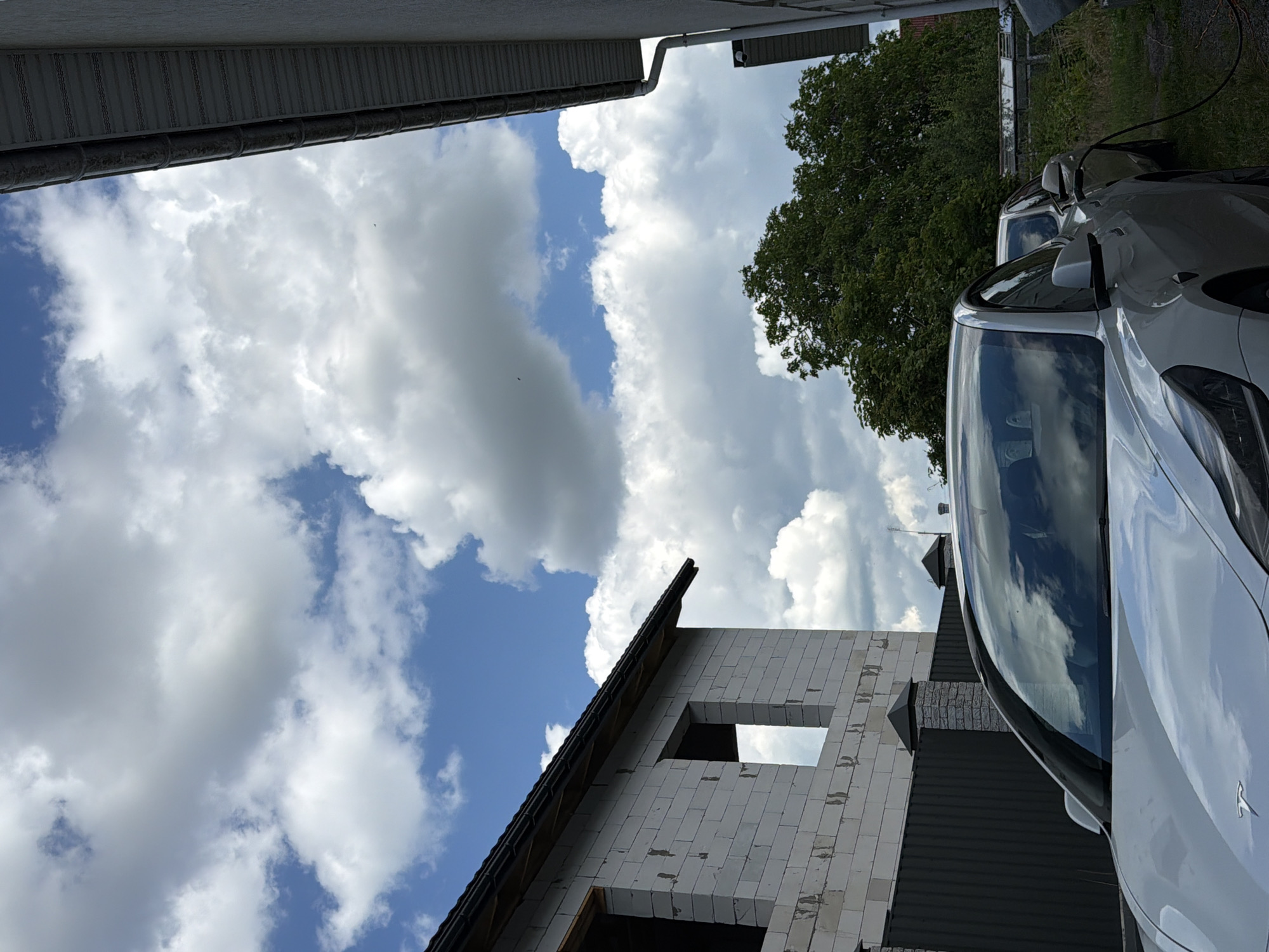 The image depicts a white Tesla electric vehicle being charged in the yard of a residential property in Bila Tserkva, Ukraine. The car is parked in front of a partially constructed building made of white cinder blocks, with visible window openings. A dark grey corrugated metal fence with stone pillars separates the property from the street. Behind the Tesla, another car is partially visible. A large, lush green tree dominates the background, with a glimpse of a greenhouse or shed-like structure behind it. The sky is filled with dramatic, puffy white clouds against a bright blue backdrop. The scene appears to be captured on a sunny day.