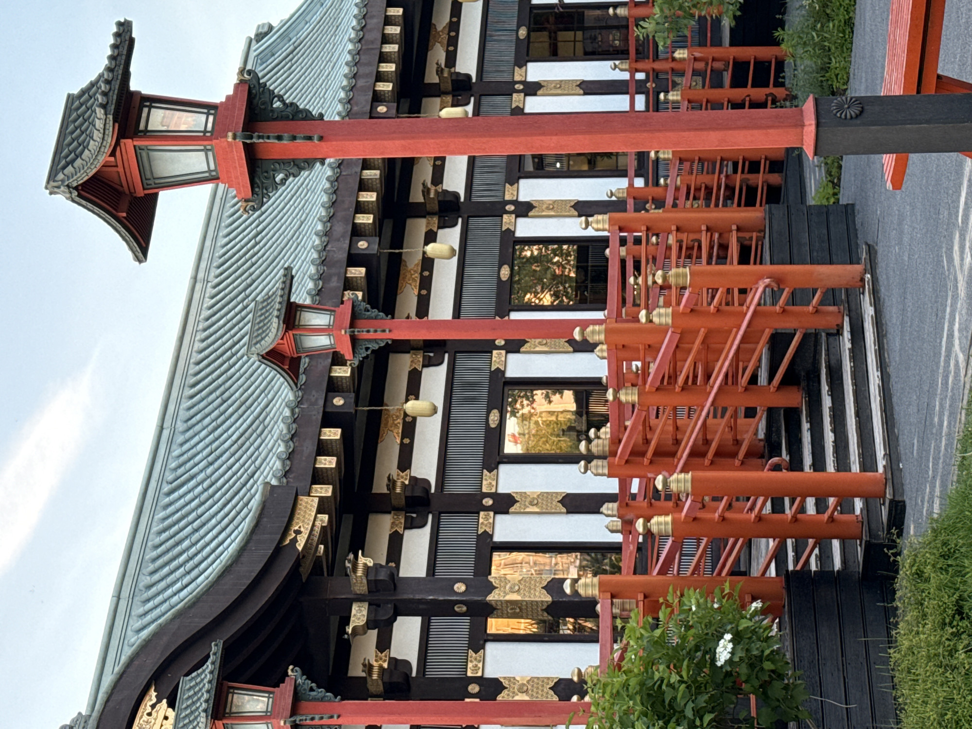 This media file features a detailed view of a traditional Japanese-style building, characterized by its distinctive architecture and vibrant colors. The prominent feature is a large, multi-layered roof with greenish-grey, textured tiles and gently curving eaves. Elaborate golden decorative elements are visible along the edges of the roof and facade.
The building's exterior consists of dark wooden beams and panels contrasting with white sections, some of which feature horizontal slatted details. Several windows reflect ambient light and hints of green foliage. Hanging spherical lanterns are intermittently placed along the building's facade.
In front of the building, a series of bright red wooden structures create a barrier or decorative fence. These structures consist of multiple horizontal and vertical bars, often topped with small, golden, spherical finials. One notable black wooden post among these red structures features a dark circular emblem, possibly a stylized chrysanthemum crest.
The scene appears to be outdoors during daytime, under a clear, bright sky. Green bushes and grass are visible at the base of the structures and along the ground, suggesting a garden or landscaped area. No people or specific activities are depicted, giving the scene a serene and static quality. There is no visible text in the image.