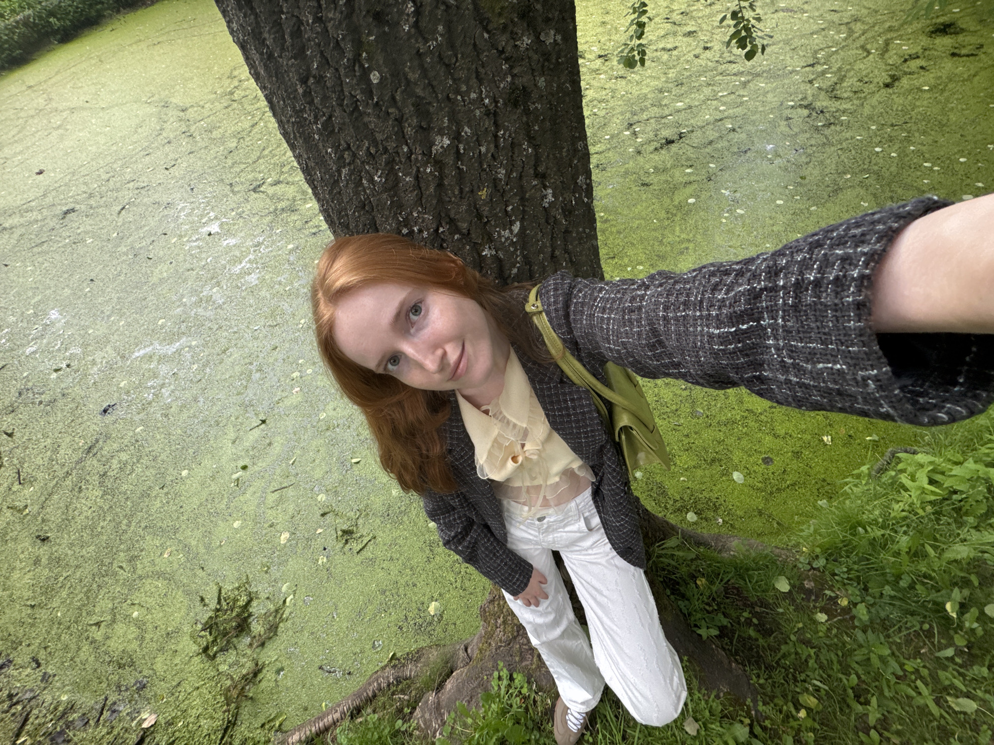 A young woman with red hair is taking a selfie in a park, with a pond covered in green algae in the background. She is wearing a grey tweed jacket, a sheer yellow blouse, and white pants. She is sitting on the ground next to a tree, with her legs crossed. The pond appears to be stagnant and filled with duckweed. The scene is shot from a high angle, looking down on the woman and the pond. The image does not contain any specific landmarks or details that would allow for definitive identification of the location in Saint Petersburg, Russia.