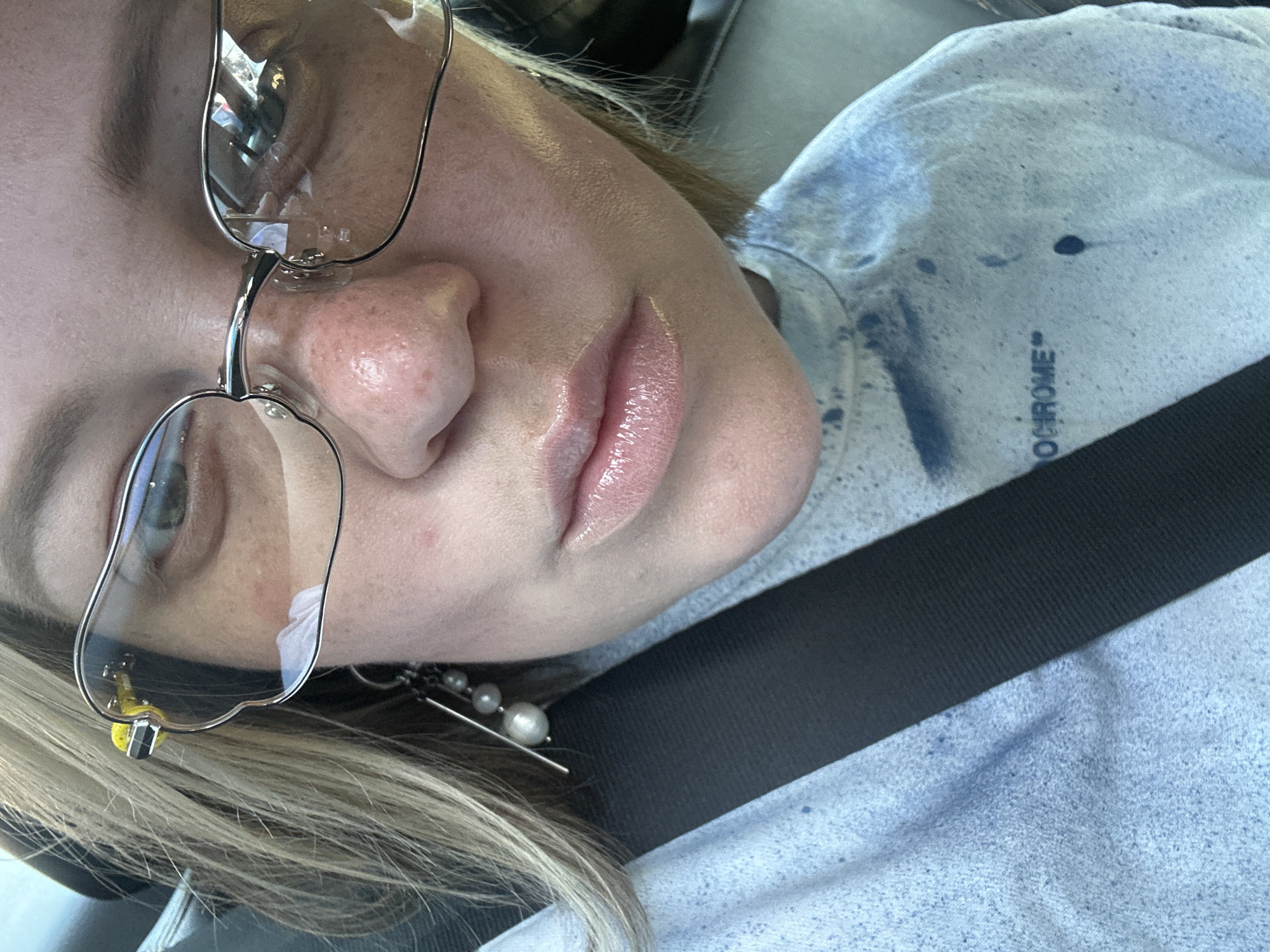 A woman with blonde hair and light skin, wearing glasses and a light blue t-shirt, is shown in a close-up shot. She has a light dusting of freckles across her nose and cheeks, and her lips are glossy and light pink. She is wearing an earring with pearls. A black seatbelt is visible across her chest. The t-shirt has a blue spray paint-like pattern and text that reads "OCHROME". The background appears to be the interior of a vehicle, with hints of a car's exterior visible in the reflection of her glasses. There is no discernible location in Moscow, Russia, as the image only provides a close-up of the woman.
