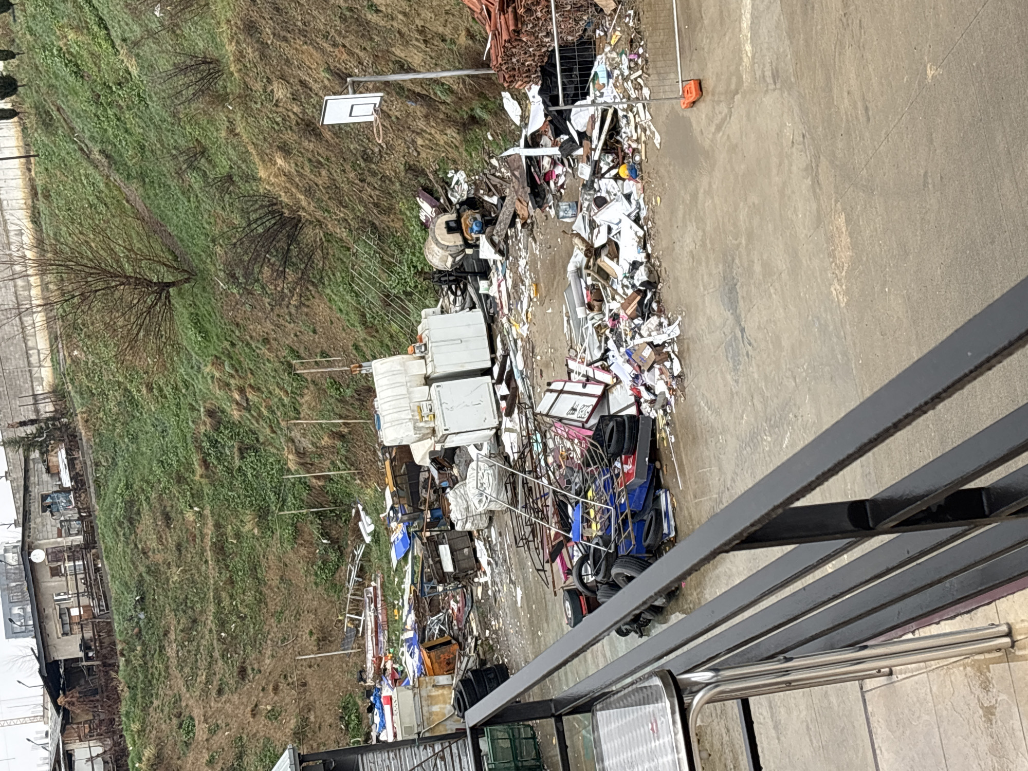 The image presents an outdoor scene in Tbilisi, Georgia, depicting a substantial and varied collection of discarded items at the base of a steep, vegetated hillside. The viewpoint is from an elevated position, indicated by a black metal railing with five horizontal bars extending diagonally across the lower foreground, partially obscuring a silver-legged table on a tiled surface.
The midground is dominated by a large, wet concrete area serving as an unofficial dump site. Visible refuse includes multiple white refrigerators or freezers, a cement mixer, numerous vehicle tires (some stacked, some dispersed), large white industrial bags, various pieces of rusted scrap metal, fragments of wood, plastic containers, discarded furniture components, and general household waste consisting of paper, plastic, and fabric remnants. On the right side of the concrete area, a freestanding basketball hoop with a white backboard and orange rim is present, alongside a stack of terracotta roof tiles and an orange traffic barrier base.
The background features a steep hillside covered with sparse green vegetation, patches of exposed earth, and several bare deciduous trees and shrubs, suggesting a non-summer season. Towards the crest of the hill, multiple rudimentary, single-story structures with fences are visible, one featuring a small satellite dish. The sky above is overcast. No individuals are present within the depicted scene.