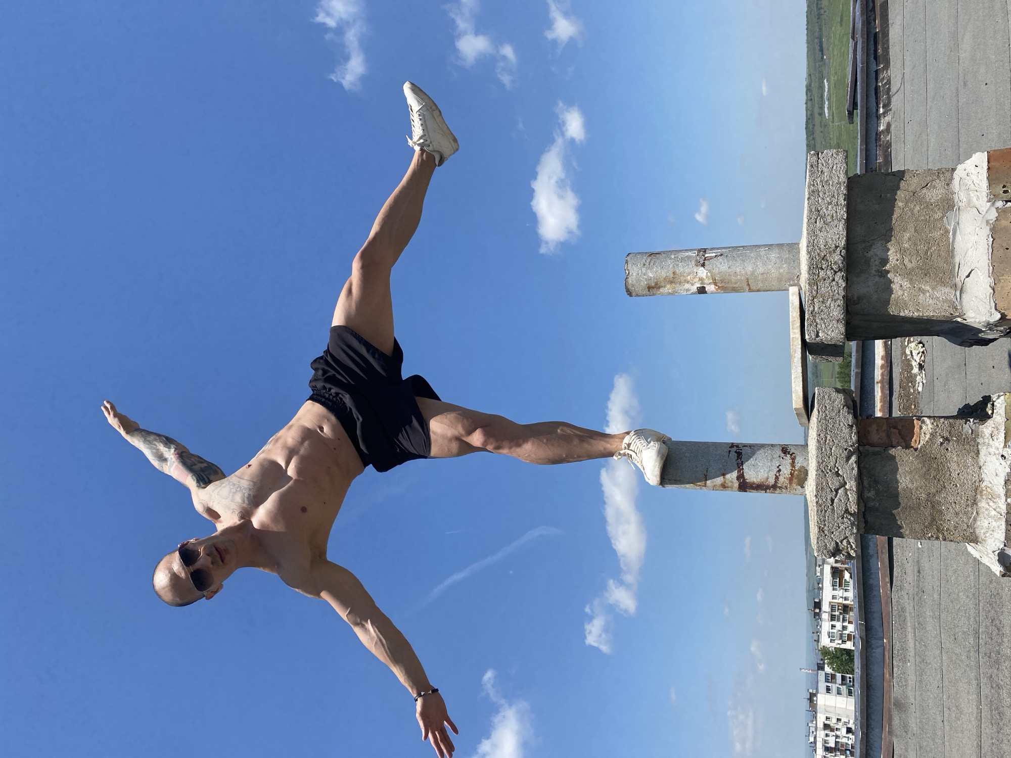 A bald man with sunglasses and arm tattoos is performing a balancing act on a concrete structure with metal rods in an outdoor setting, presumably the city of Pleven, Bulgaria. The man is shirtless, wearing black shorts and white sneakers, with one foot on each of the metal rods, arms outstretched. The background consists of a blue sky with scattered clouds and a cityscape on the horizon. The structure appears to be located on a rooftop.