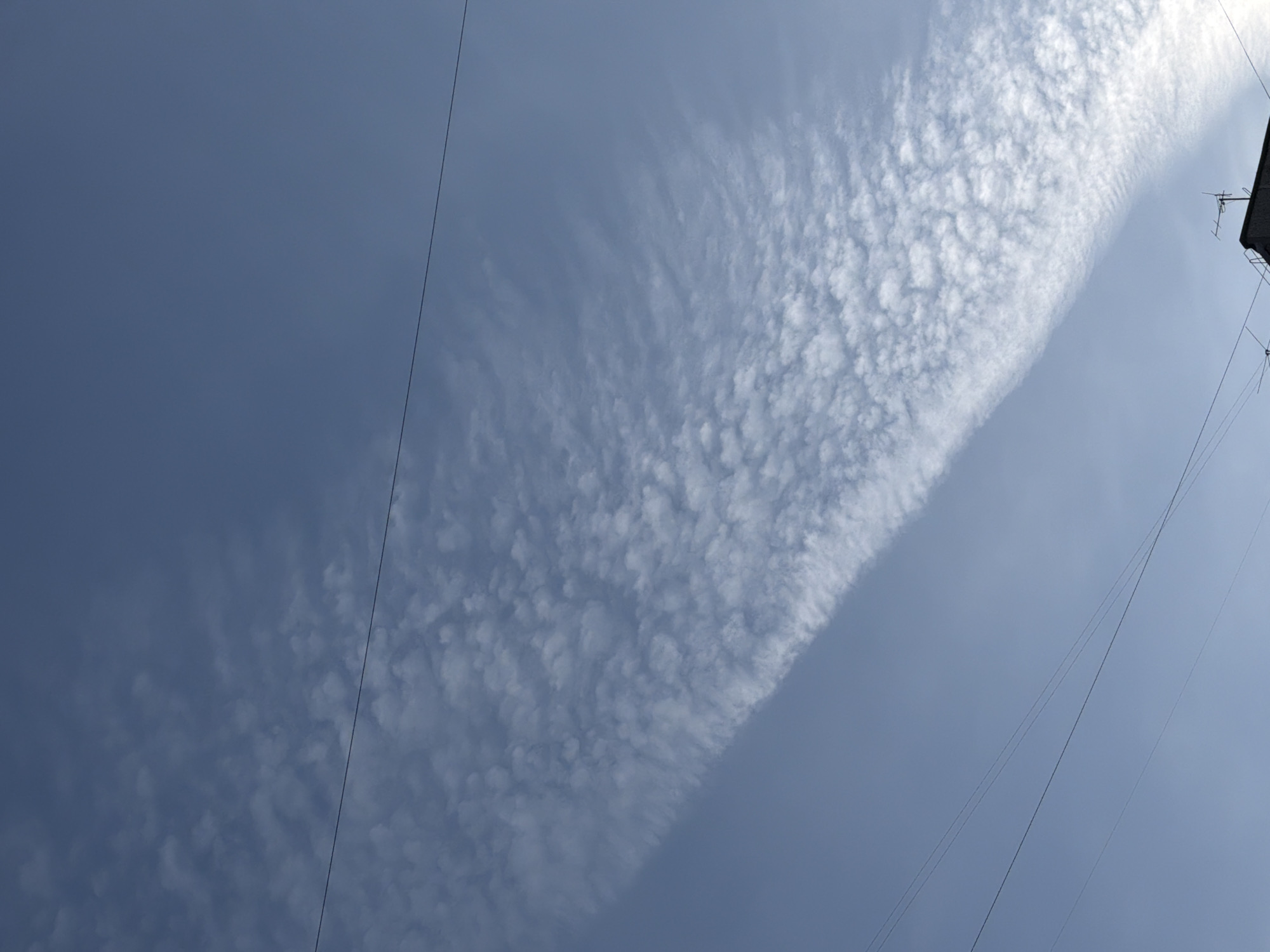 The image displays a clear to partly cloudy sky dominated by a striking diagonal band of textured, white clouds. This cloud formation, stretching from the lower-left to the upper-right of the frame, exhibits a rippled or mackerel-like pattern, characteristic of altocumulus or cirrocumulus clouds. To the left of this prominent cloud band, the sky is a deeper, more uniform shade of blue, while the area around the clouds is a lighter, brighter blue, indicating a sunny day.
Several thin, dark lines, identifiable as power lines or communication cables, traverse the sky diagonally, adding a man-made element to the natural scene. One prominent wire runs along the far-left edge, and multiple other wires are visible in the central and right portions of the frame. In the upper-right corner, a small section of a building's roofline or upper structure is visible, featuring an antenna or aerial, suggesting an urban or suburban setting.
The image captures a bright daylight scene with no people or specific activities depicted. There is no visible text.