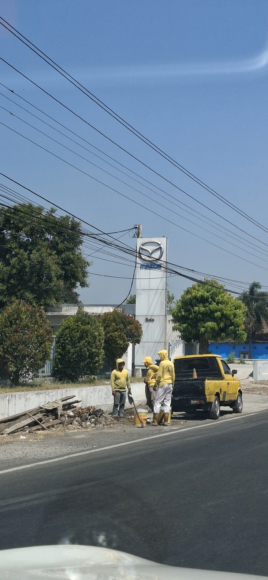 The image depicts a street scene in Ngadirejo, Indonesia, under a clear blue sky. In the background, a Mazda dealership building is visible, with its distinctive logo prominently displayed on a tall white pylon. The dealership is flanked by manicured trees and bushes. A yellow pickup truck is parked on the side of the road, its bed empty except for an orange traffic cone. Three workers, all wearing yellow hooded jackets and pants, and yellow boots, are gathered near the truck. One worker, wearing blue jeans and a yellow jacket, holds a shovel. Another worker, also in yellow, is bent over, possibly working on the ground. A third worker in yellow stands facing away from the camera. A pile of debris, including wood scraps and rubble, lies on the side of the road next to the workers. Power lines crisscross the upper portion of the image, creating a network against the sky.