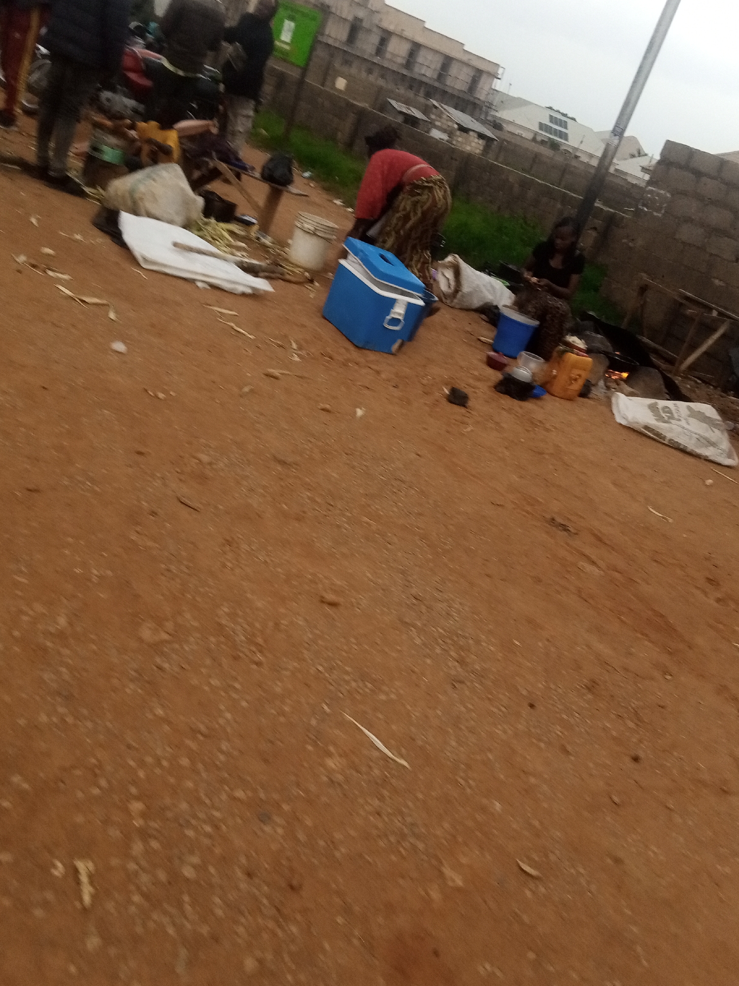 In a dusty, outdoor urban setting, a group of individuals are gathered around a collection of objects. A person wearing a red shirt and a patterned skirt is bent over, engaged in an activity. Beside them, a blue cooler sits on the ground. Further to the right, a young woman in a dark top and patterned skirt is seated, also occupied with something. The ground is covered in reddish-brown dirt, with scattered debris, including what appears to be straw or dried vegetation. In the background, various buildings, some with multiple stories and brick construction, line the area, suggesting a developed urban environment. A tall pole stands prominently in the mid-ground. Several containers, including a yellow jerrycan, blue buckets, and a white sack, are visible among the belongings. The overall atmosphere is one of informal activity in a somewhat disheveled urban space.