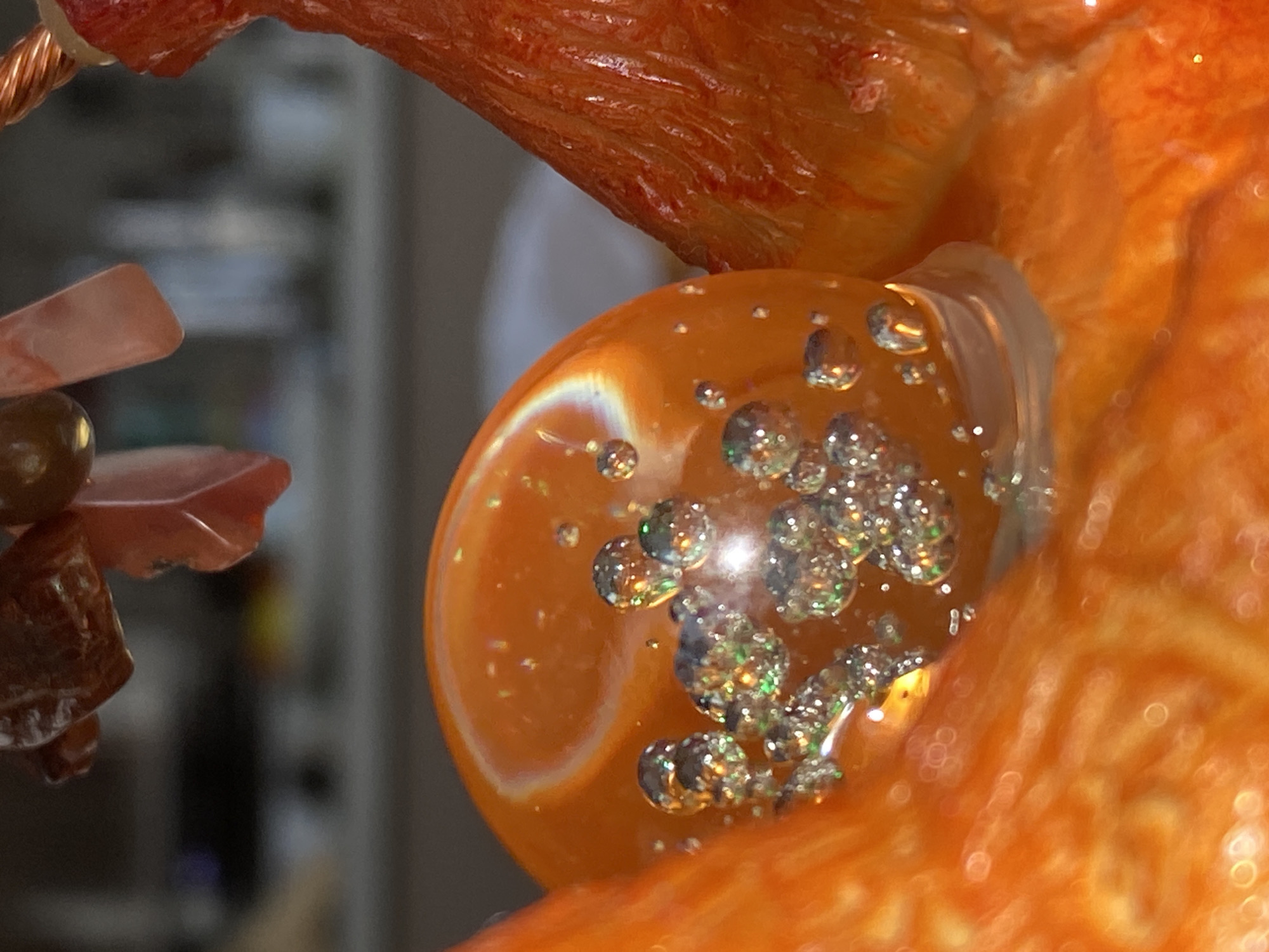 An extreme close-up shot features a vibrant, translucent orange object, likely made of resin or glass, occupying the central and right portions of the frame. Within this smooth, glossy object are numerous small, iridescent particles or bubbles, glittering with reflections of light that display hints of green, silver, and gold. A pronounced white streak of light refracts through the orange material on the left side of the main object, indicating its rounded surface.
To the upper left, a tightly coiled copper wire with a metallic sheen is partially visible, appearing to connect to or be part of the larger orange structure. Below this wire and to the far left, several smaller, reddish-brown objects, possibly polished stones or beads with a more irregular, textured surface, are partially seen.
The background is heavily blurred and out of focus, appearing as indistinct shapes and muted colors, suggesting an indoor setting. The shallow depth of field emphasizes the intricate details and light play on the central orange element. There is no discernible activity taking place, and no people or visible text are present. The lighting appears to be bright and direct, creating strong highlights and sparkle. The overall impression is that of a decorative item, possibly a piece of jewelry or a sculpture, such as a wire-wrapped stone "tree." No specific cues within the image indicate the location of Фоки, Russia.