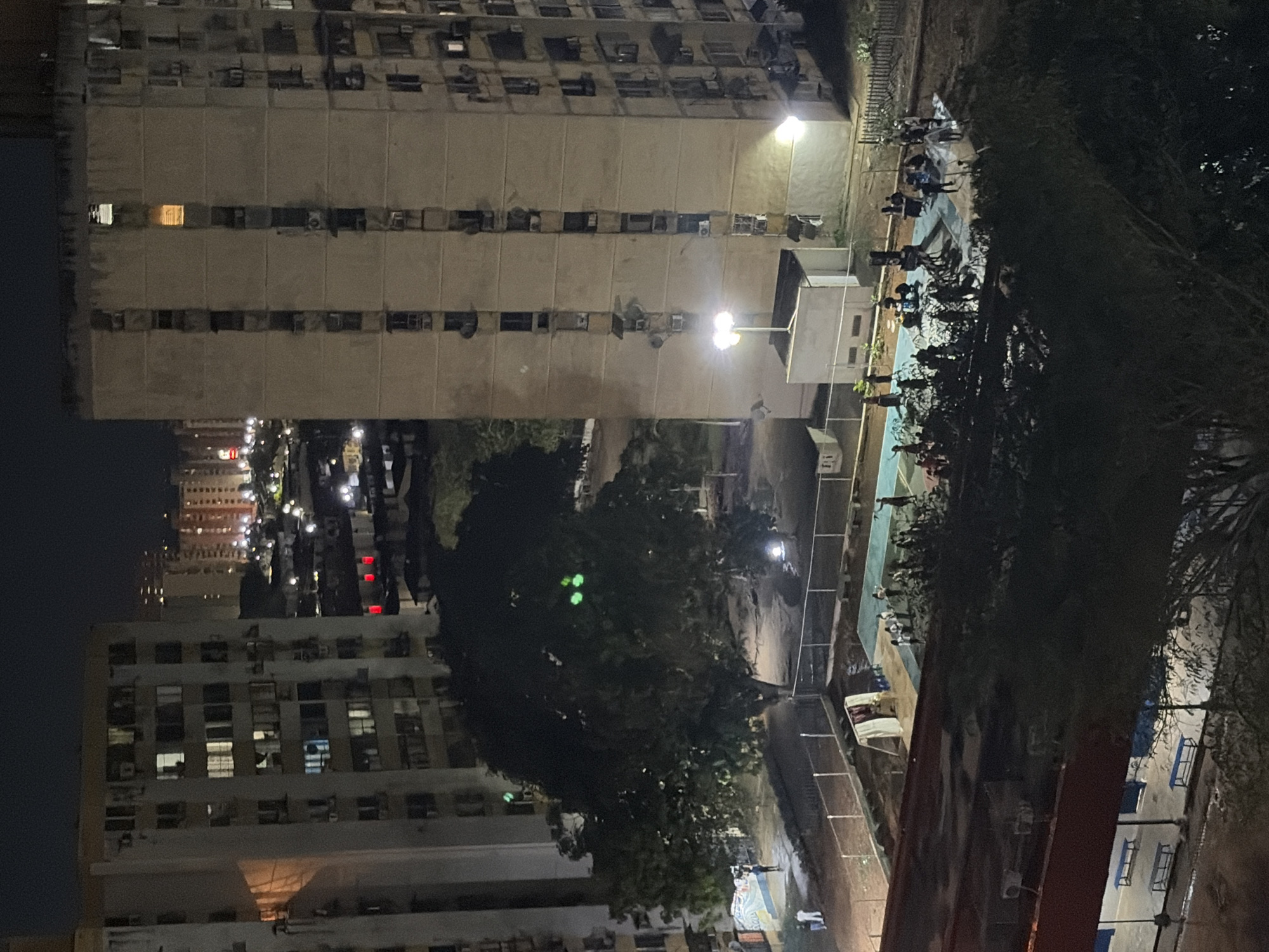 An eye-level, nighttime image shows a high-density urban environment in Ciudad Guayana, Venezuela. The scene primarily features a cluster of tall, aged apartment buildings with numerous windows and air conditioning units. The buildings are densely packed together and display signs of weathering. In the midground, a cluster of trees divides the composition. A recreational area is visible, including a basketball court occupied by a group of people. Street lights illuminate the area. The image is somewhat dark and grainy, giving a sense of urban density and nighttime activity.