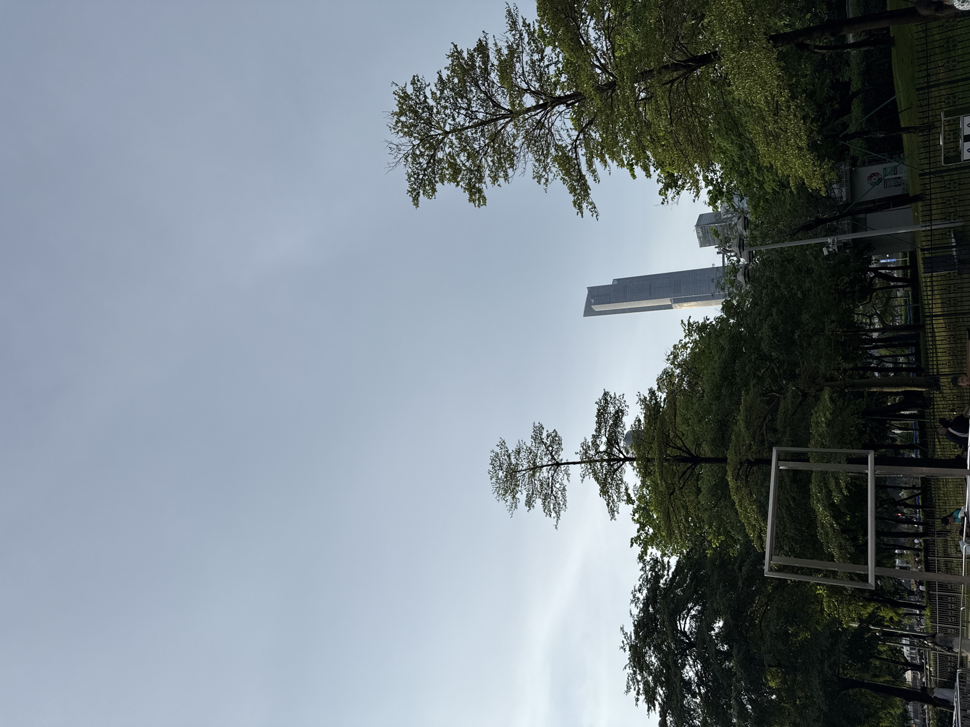 In Haizhu District, China, a clear sky above a park features lush green trees framing a view of modern skyscrapers. A prominent tall, slender skyscraper with a distinctive angled top dominates the background, flanked by shorter buildings. In the foreground, a large, abstract metal square sculpture is mounted on posts. A black metal fence separates the park area from a pathway or road where several indistinct figures are visible. The scene depicts a blend of urban infrastructure and green space.